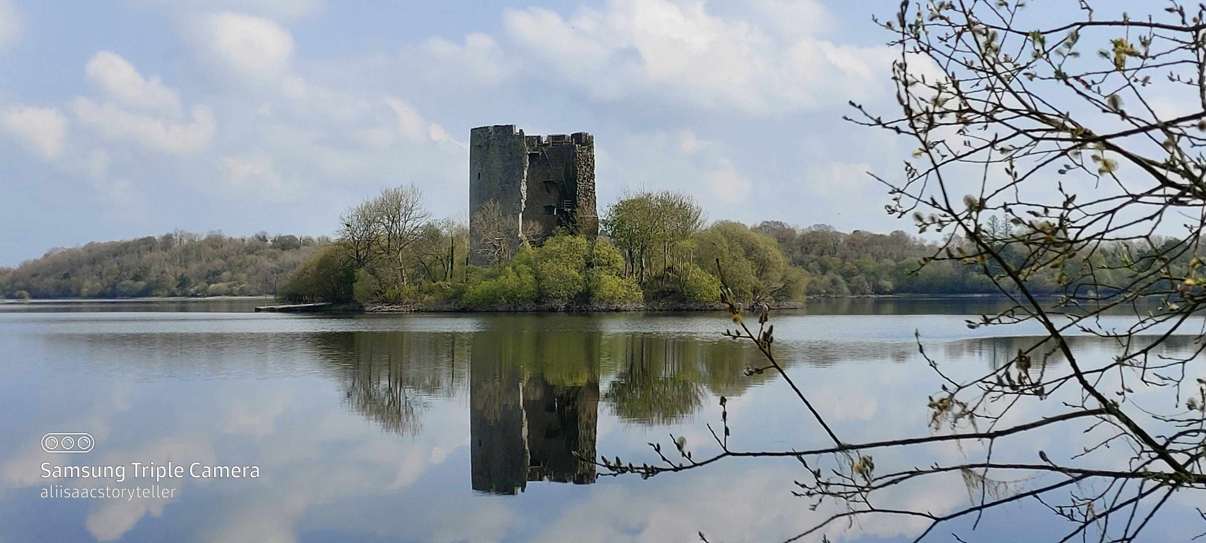 The ruined face of a round tower-house style castle appears to rise out of the still waters of a lake, its base shrouded with trees and shrubs. The cloud-studded sky is perfectly reflected in the lake, the water is still as a mirror.