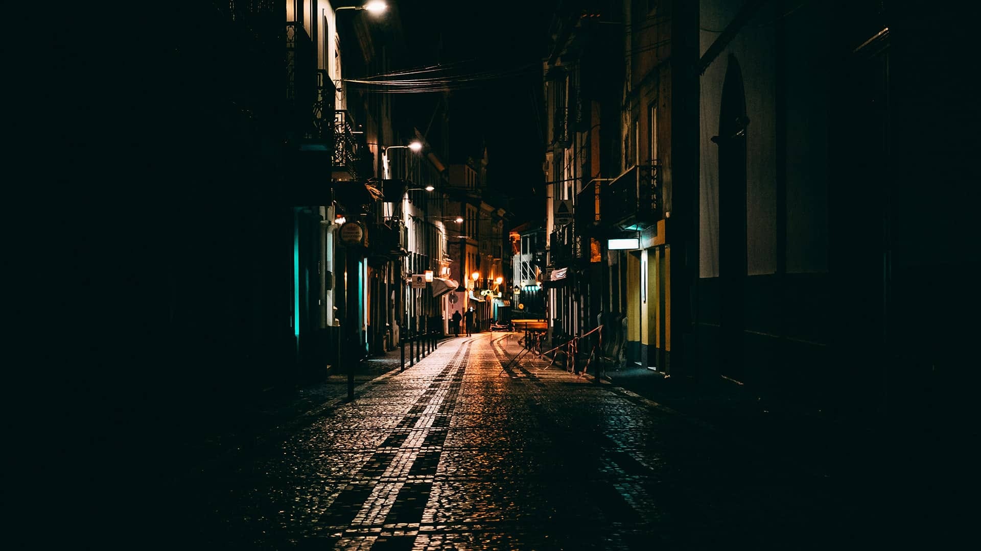 A wet cobblestone street at night reflecting the glow of streetlights, with buildings on each side deep in the shadows.