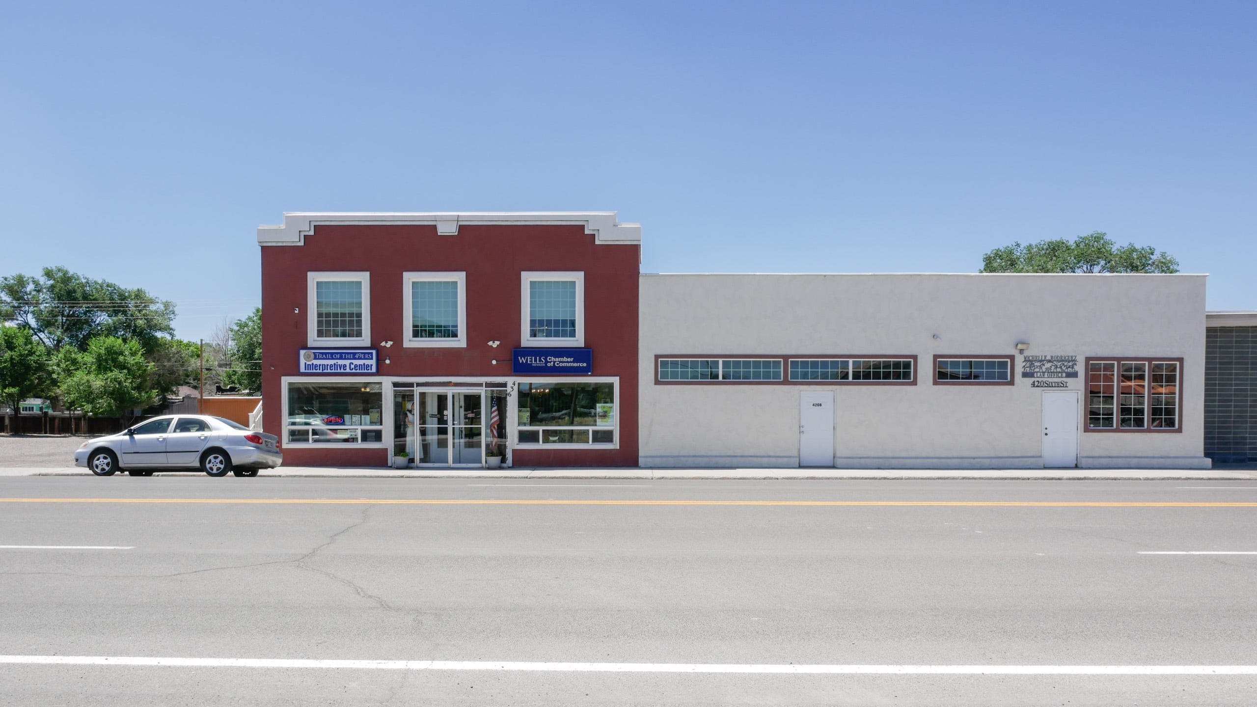 Photo looking across an empty street at two modest buildings. To the right is a low, one and a half-story, flat roofed, white building with two doors (also white), three taller windows to the right, and long, narrow, windows along the left. The building is a simple rectangle without any adornments. To the left is a two story red brick building with white trim with big plate glass windows on either side of the glass door entrance on the first floor and three large windows on the third floor. One sign on the left of the building says, "Trail of the 49ers Interpretive Center" while the sign on the right says, "WELLS Chamber of Commerce" To the left of the building, parked on the street, is a late model Toyota compact sedan, silver. Some green trees can be seen on the left and above the white building to the right.