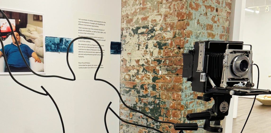 An art exhibit created by Rachel Handlin featuring a large vintage camera on a tripod, a wireframe outline of a human figure, and a photo displayed on the wall with descriptive text.