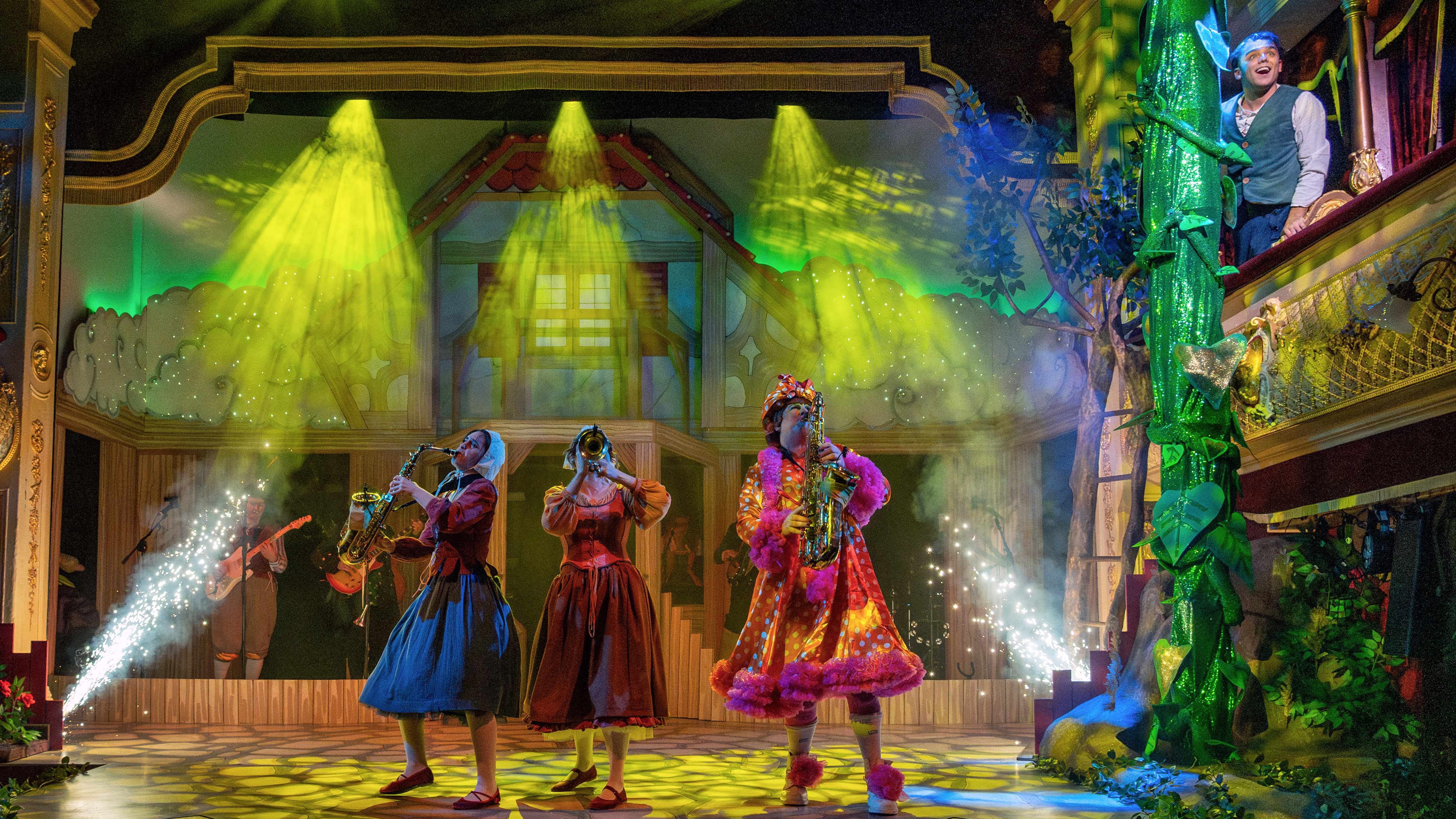 Three pantomime characters play musical instruments onstage whilst Jack looks down from a box next to an enormous green beanstalk. The old-fashioned proscenium -arch stage is suffused with green light.