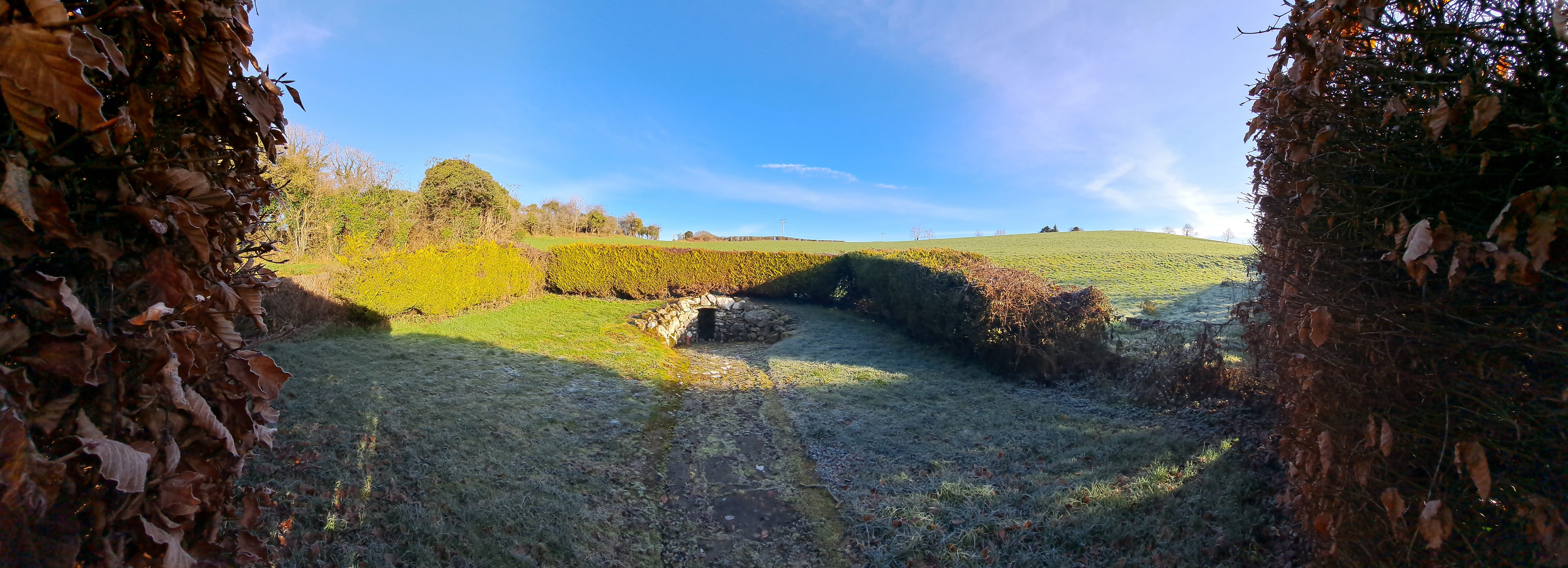 the arch of Brigid's well is glimpsed between a gab in the hedge, a flagged path leads up to the stone walled entrance, which is a small rectangular dark mouth. The site is enclsed by dense hedges, a lovely rolling meadow can be seen beyond, the sky is bright blue with some wispy clouds, there is frost on the grass.