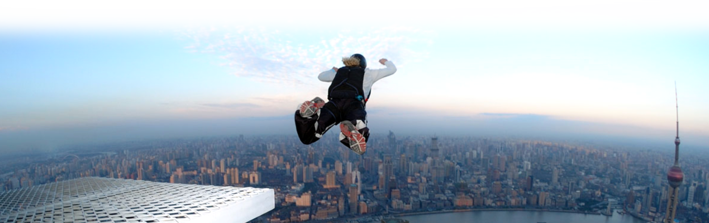 A base jumper jumping off a building with a big city skyline in the back, just before sunset.