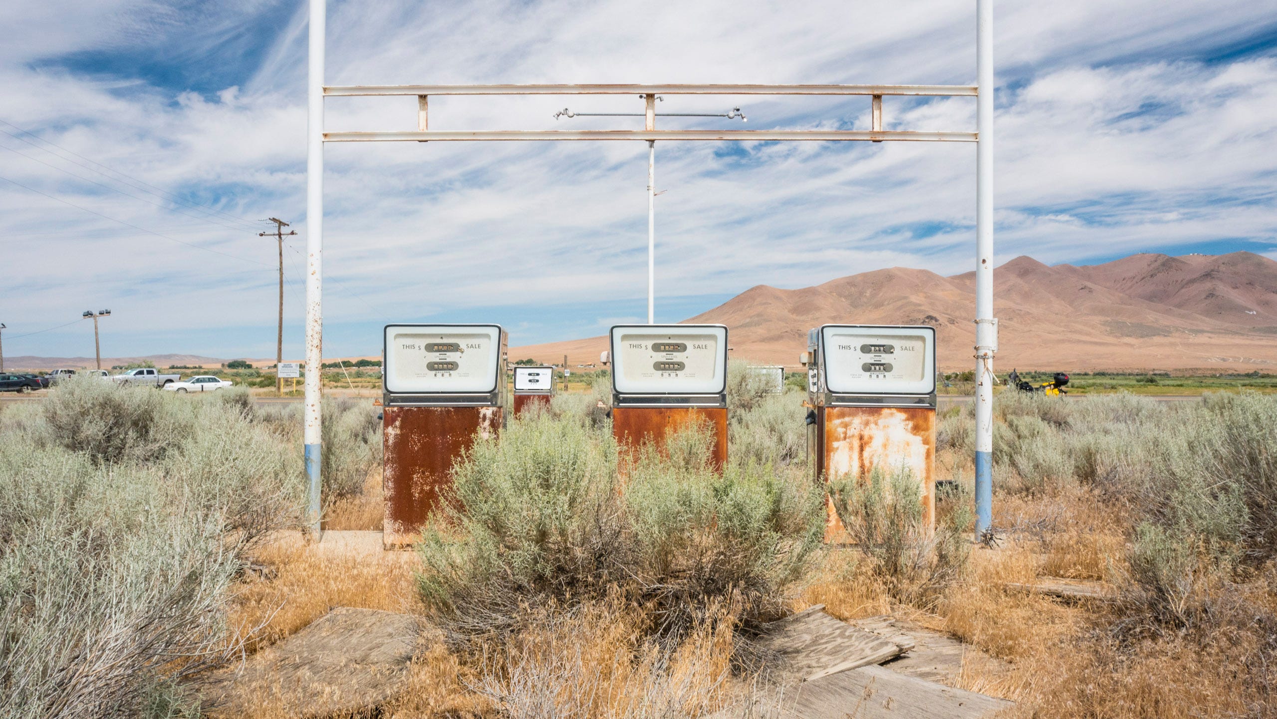 Color photograph of an old, long-abandoned mid-20th century gas station. In the center of the photo are three rectangular gasoline pumps, their lower portions rusty from prolonged exposure to the weather. An overhead gantry that may have held an awning is above the pumps but there is no awning. The paved area where cars would pull up alongside the pumps has been overrun with sagebrush and other desert scrub brush. In the distance are jagged and naked mountains and blue sky streaked with clouds.