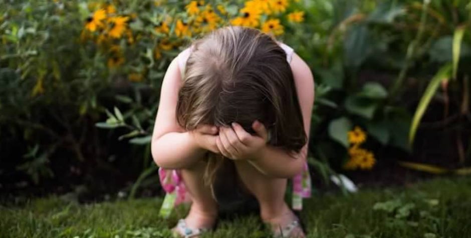 a 2012 photo of Penny crouching in the grass in a garden, covering her face with both hands. Behind her, there is a backdrop of vibrant yellow flowers