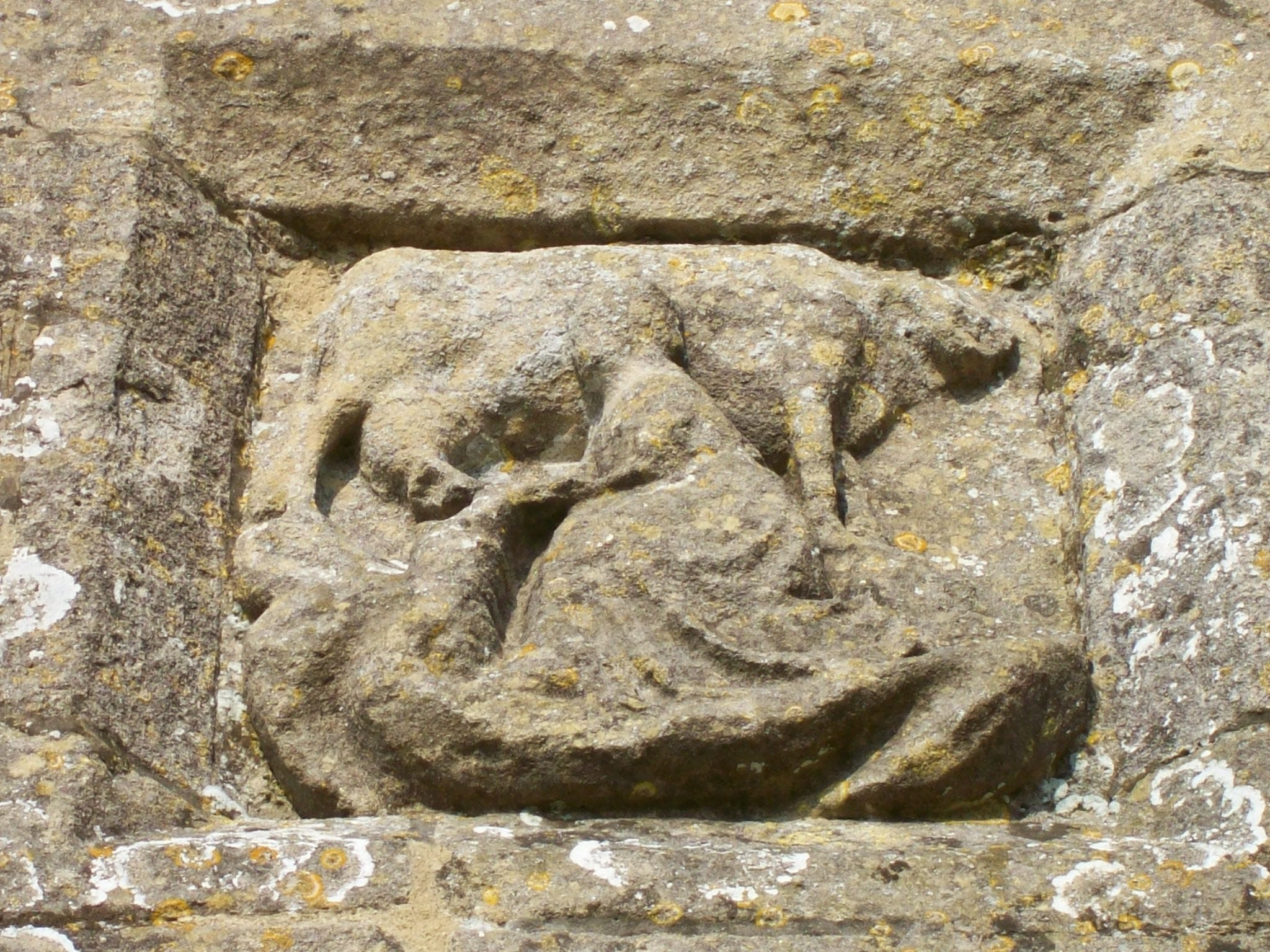 Carving of cow being milked by a woman in long skirt, the stone is sandy in colour and spotted with patches of green and white lichen.