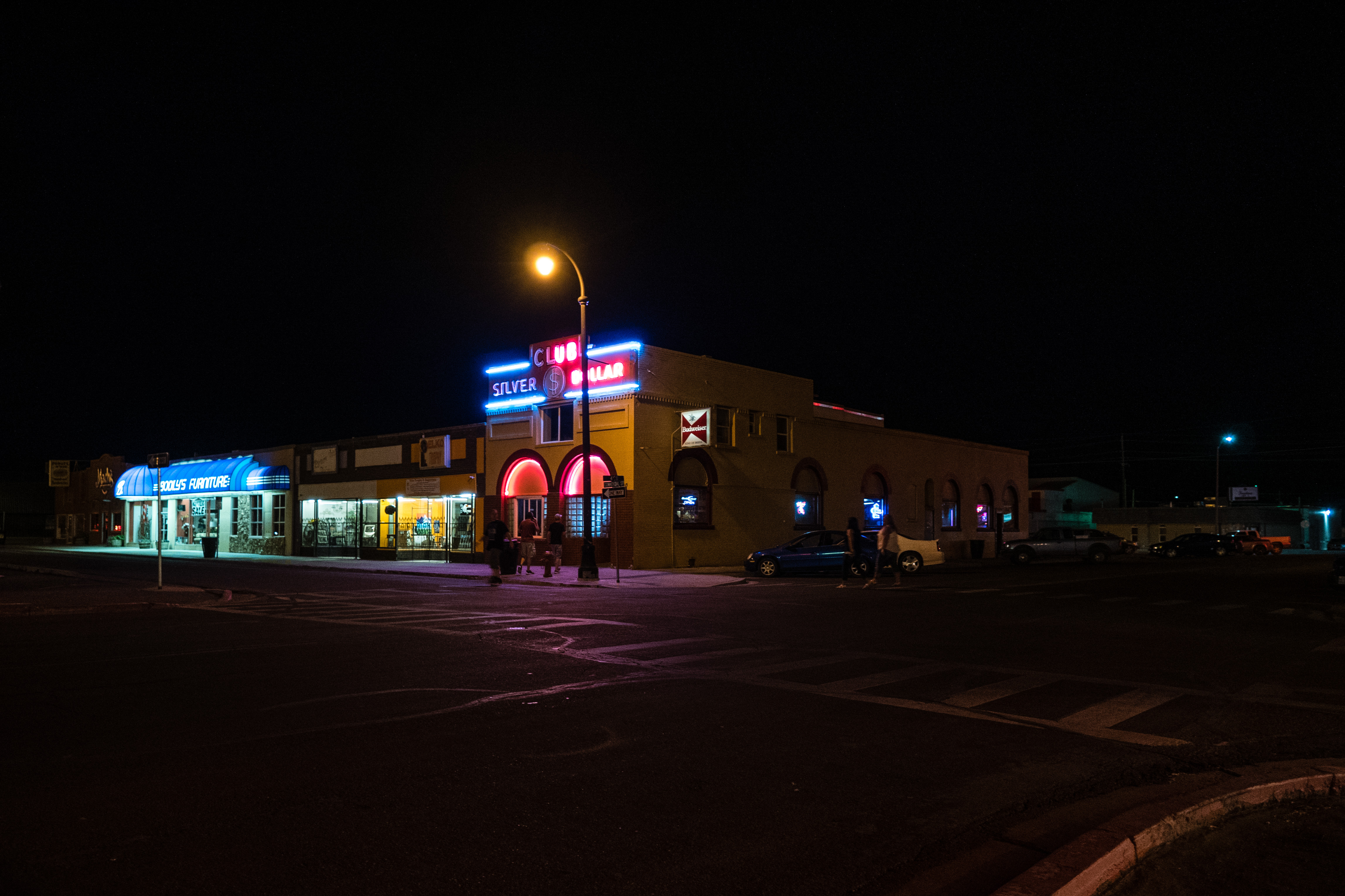 Evening photograph of a street corner in Elko, Nevada. On the corner is a long, low, older building, pale yellow with arched windows. At the top of the facade, in neon, is "SILVER DOLLAR CLUB" in red and blue neon. The letters "C" and "L" of the word "CLUB" are unlit, possibly broken. Below is the entrance to the club, with neon outlining the arched above the entrance and the arch above the main window. To the side, are additional windows with neon signs of alcohol brands. To the left are additional local shops with their display windows lit but they are otherwise closed. One of the stores has a blue awning with the word "FURNITURE" clearly visible. A single street lamp stands on the corner in front of the club. The sidewalk is dimly lit by the shop lights and street lamp. To the right of the club, cars are parked in the shadows. Two people can be seen walking in front of the club. Two woman can be seen walking across the street to the club. They are blurry.