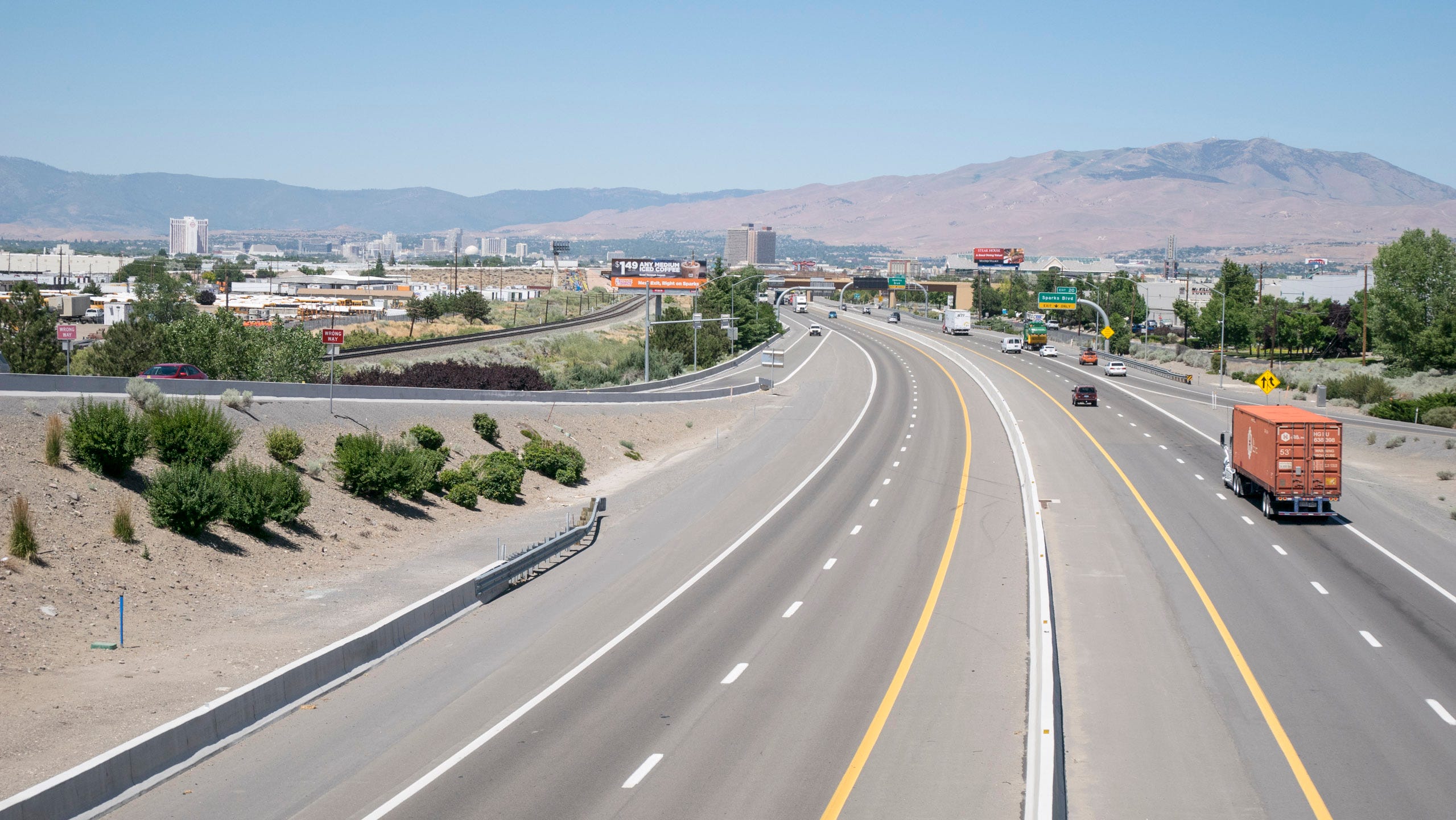 Photo of a wide interstate, two lanes in each direction with generous shoulders on each side, arcing towards downtown Reno, Nevada. There are a smattering of cars and trucks on the road and a smattering of green vegetation in dry sandy soil on either side of the road. A highway exit ramp can be seen on the left, and beyond that are railroad tracks. The road bends and its vanishing point the skyline of downtown Reno can be seen. Beyond the buildings are tall mountains on the other side of the valley. The mountain on the right is tall and sandy and pink with just a smattering of green near its rounded peak. Mountains on the left are greener and hazier.