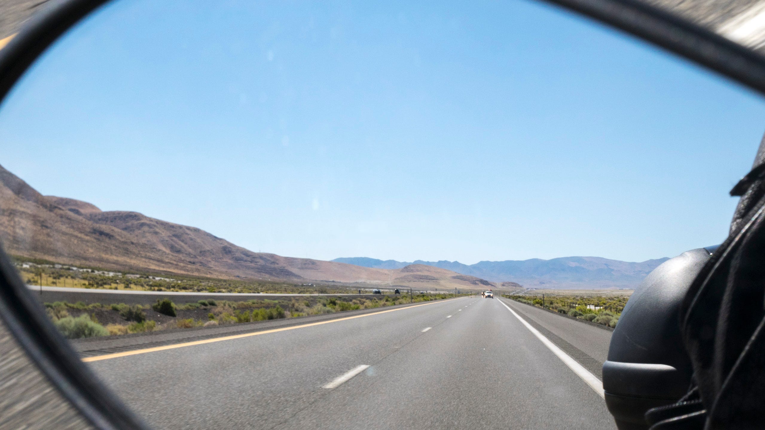 Photo of the rearview mirror of a motorcycle on a wide, modern, interstate highway in Nevada. The mirror occupies the majority of the picture frame, with just the curved edges of the mirror in the corners of the photo. In the mirror's reflection, the highway recedes arrow straight towards distant, hazy mountains. The headlights of two distant cars can also be seen. To the left, the highway median is peppered with green vegetation, and the two opposing lanes of the highway can also be seen. Beyond the opposing lanes, the land is flat with sparse, low vegetation before reaching more rugged, naked mountains.