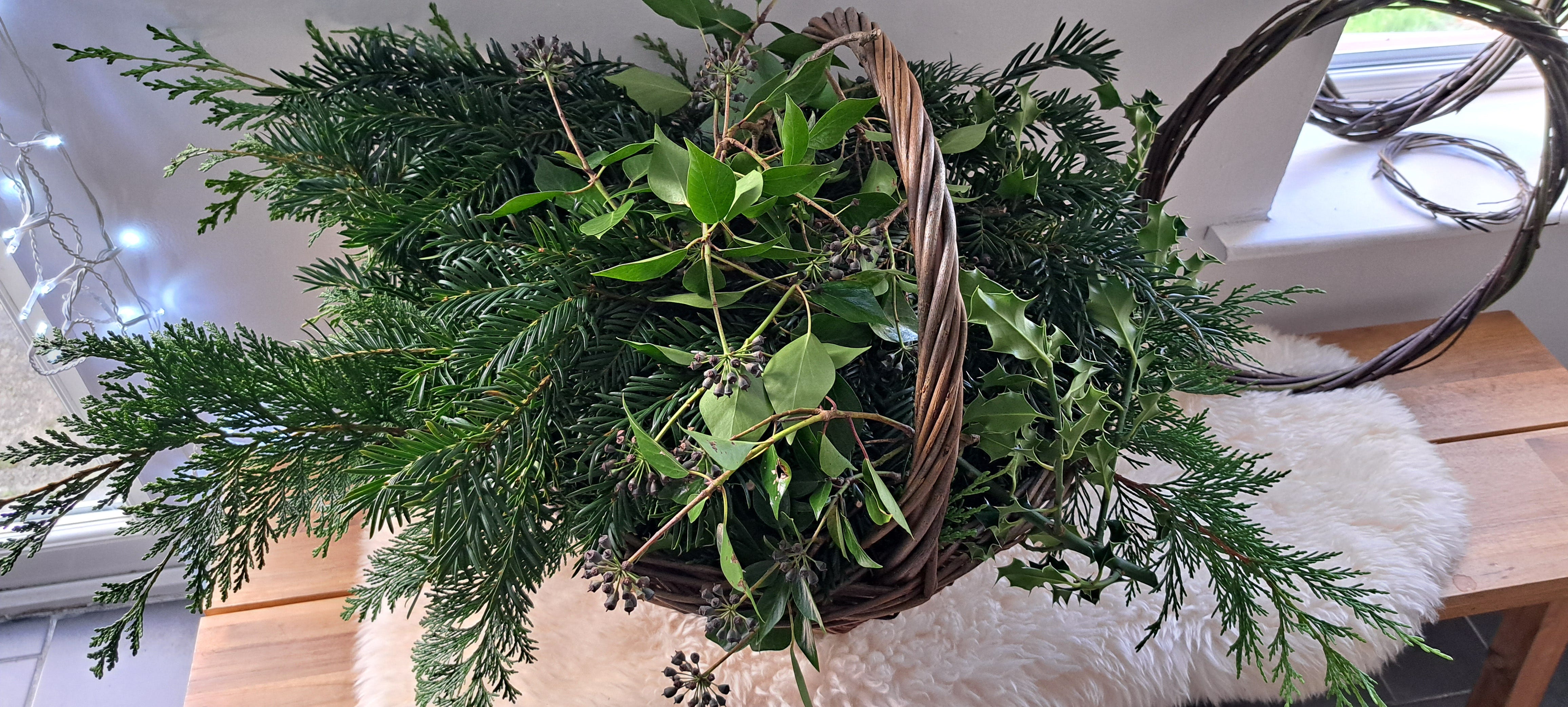 Foraged greenery in a willow basket on a bench beside two willow wreath bases.