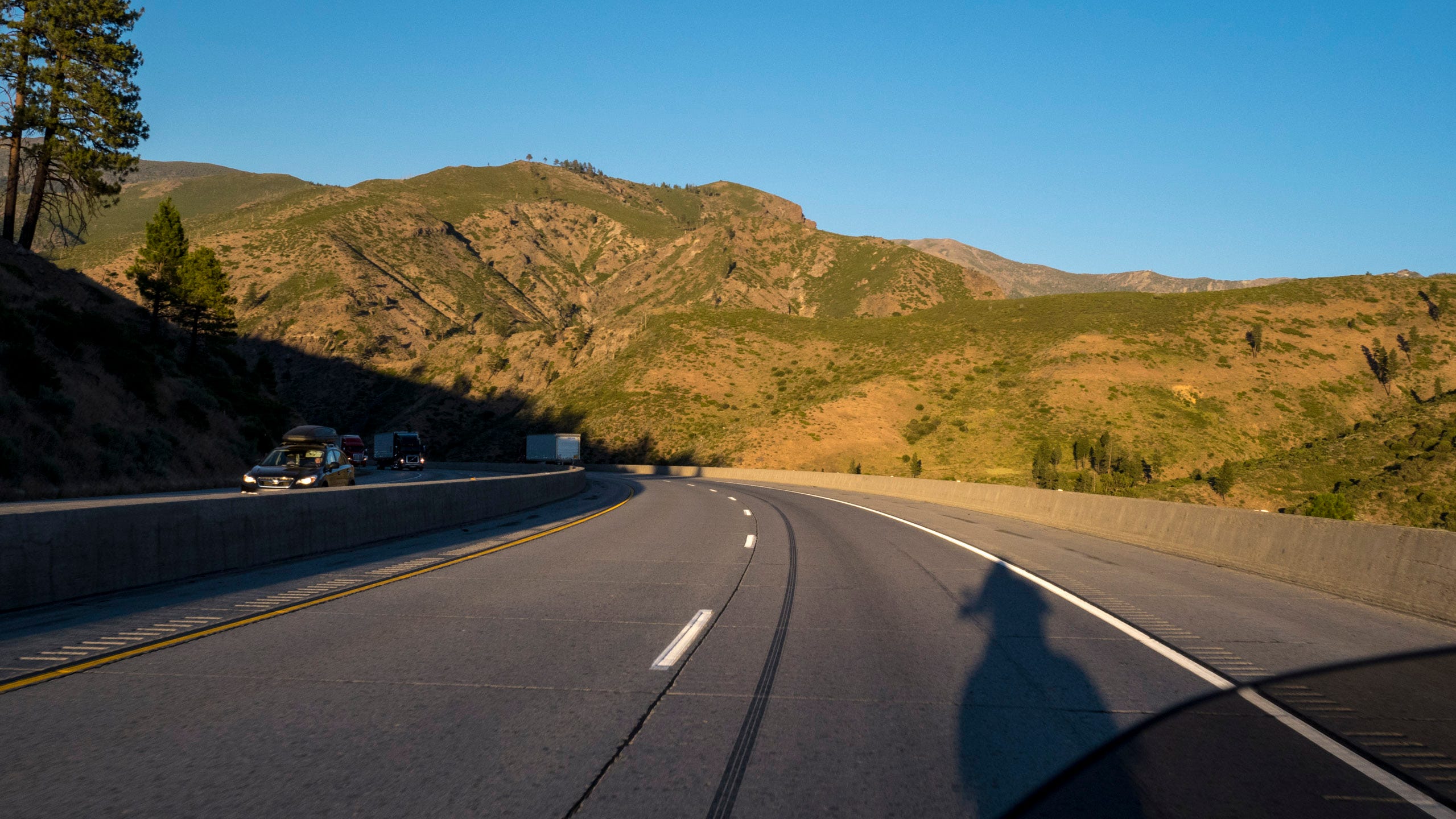 A photo in the late afternoon of a motoryclist riding down a mountain highway. The photo is taken from the motorcyclist's point of view; you can see the motorcycle's windshield in the lower right corner of the photo, the sun is behind him and you can see the shadow of him and the motorcycle on the road. The highway is descending through rugged, mountains, nearly bare but for some short scrub and grasses growing in patches and a small stand of trees at the rounded peek of the mountain directly ahead. The sun is low and casting a warm light upon the scene. There is a white 18-wheeler also descending in the distance, and a handful of vehicles climbing up the hill towards the rider and into the sun.