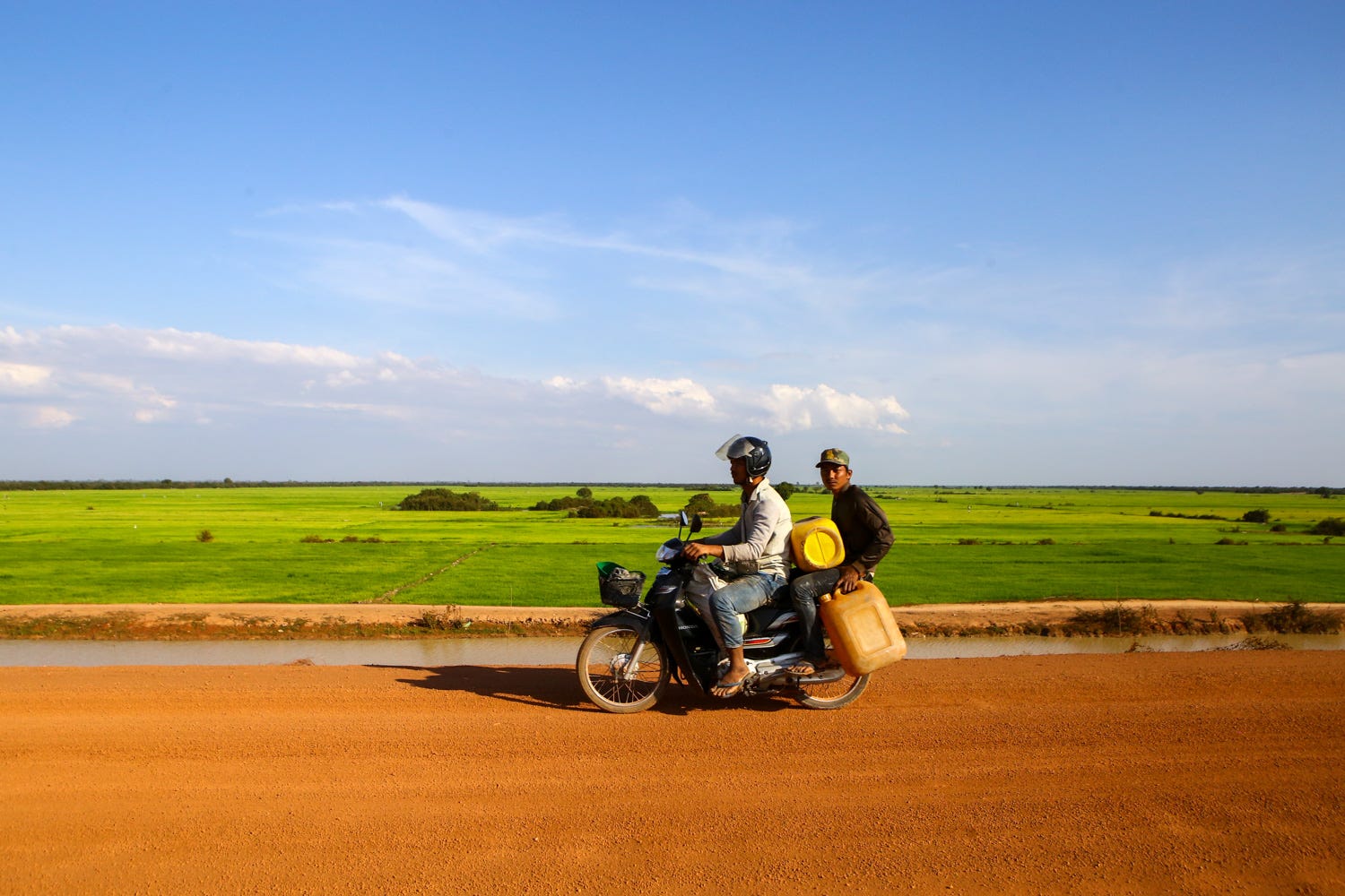 Two men are riding a motorcycle down a bright orange dirt road, one man driving and wearing a helmet and looking straight ahead. The other man is holding two gas cans and wearing a camouflaged hat and staring directly at the camera. Behind the road, green fields expand to the horizon, and the sky is a bright blue overhead.  
