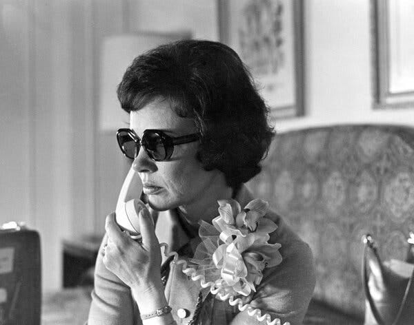 A woman holding a landline phone to her ear. She has a short haircut, is wearing dark sunglasses and is sporting an orchid corsage.