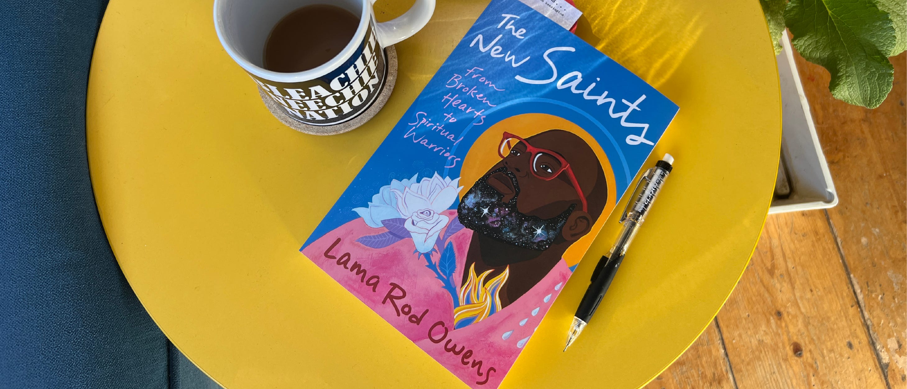 Photo of the book The New Saints sitting on a small round yellow side table. A mechanical pencil is laying to the right of the book and a mug of tea sits to the left. 