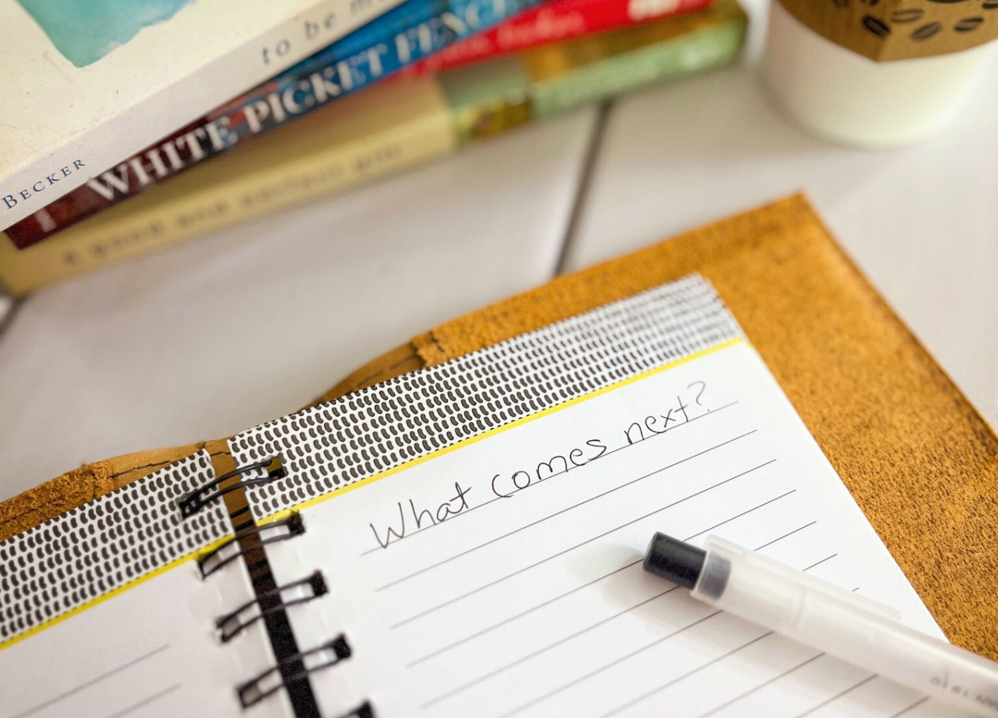 A close-up image of an open notebook with the handwritten words “What comes next?” on a lined page. A black-capped pen rests on the page. In the background, a stack of books is slightly out of focus, with one spine reading “WHITE PICKET FENCES” and another showing the author's name, “Becker.” A coffee cup with a sleeve decorated in a coffee bean pattern sits nearby. A close-up image of an open notebook with the handwritten words “What comes next?” on a lined page. A black-capped pen rests on the page. In the background, a stack of books is slightly out of focus, with one spine reading “WHITE PICKET FENCES” and another showing the author's name, “Becker.” A coffee cup with a sleeve decorated in a coffee bean pattern sits nearby.
