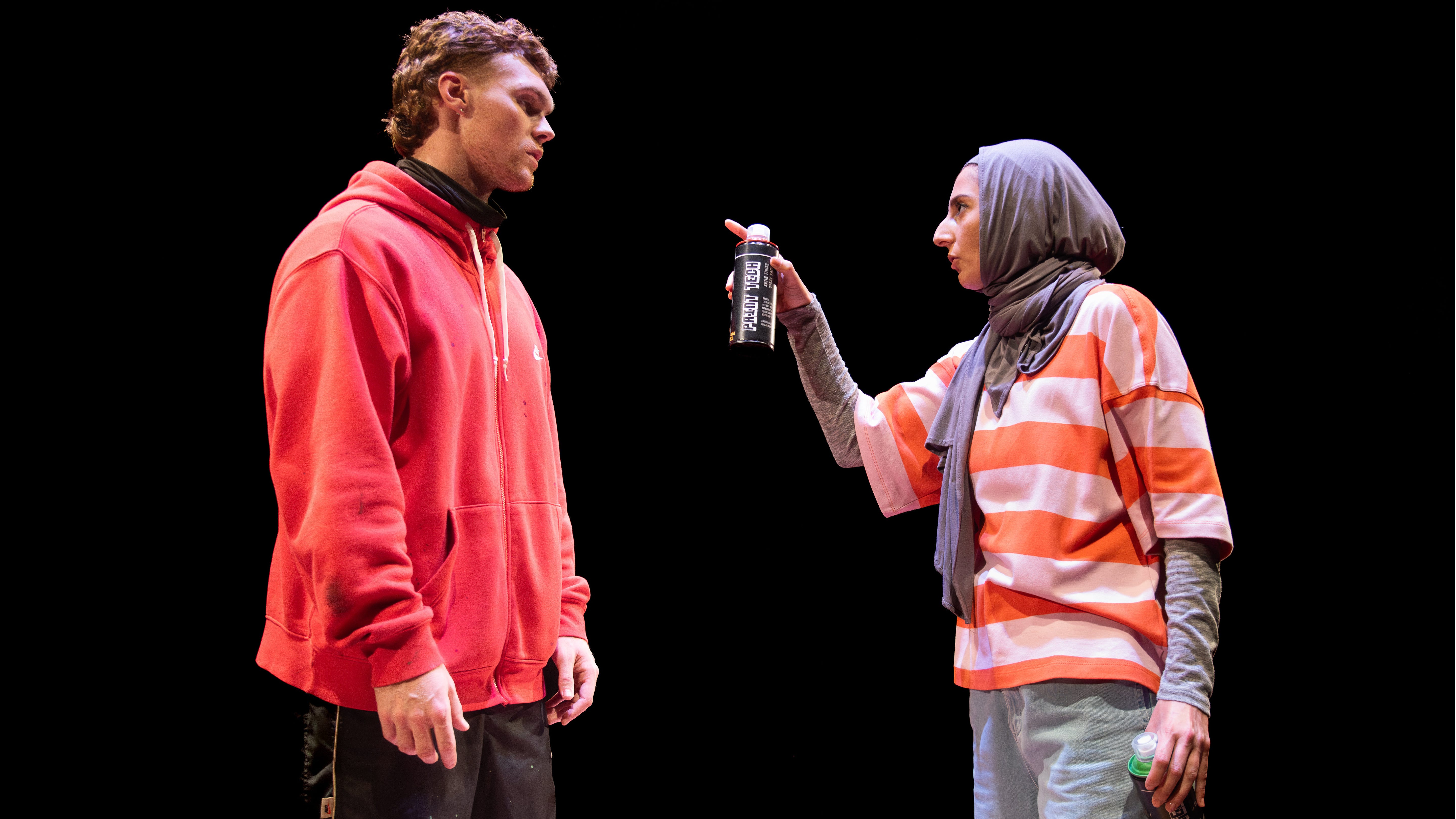 A young woman in a headscarf gestures at a man wearing a hoodie. She is holding a spray-paint can and looks angry.