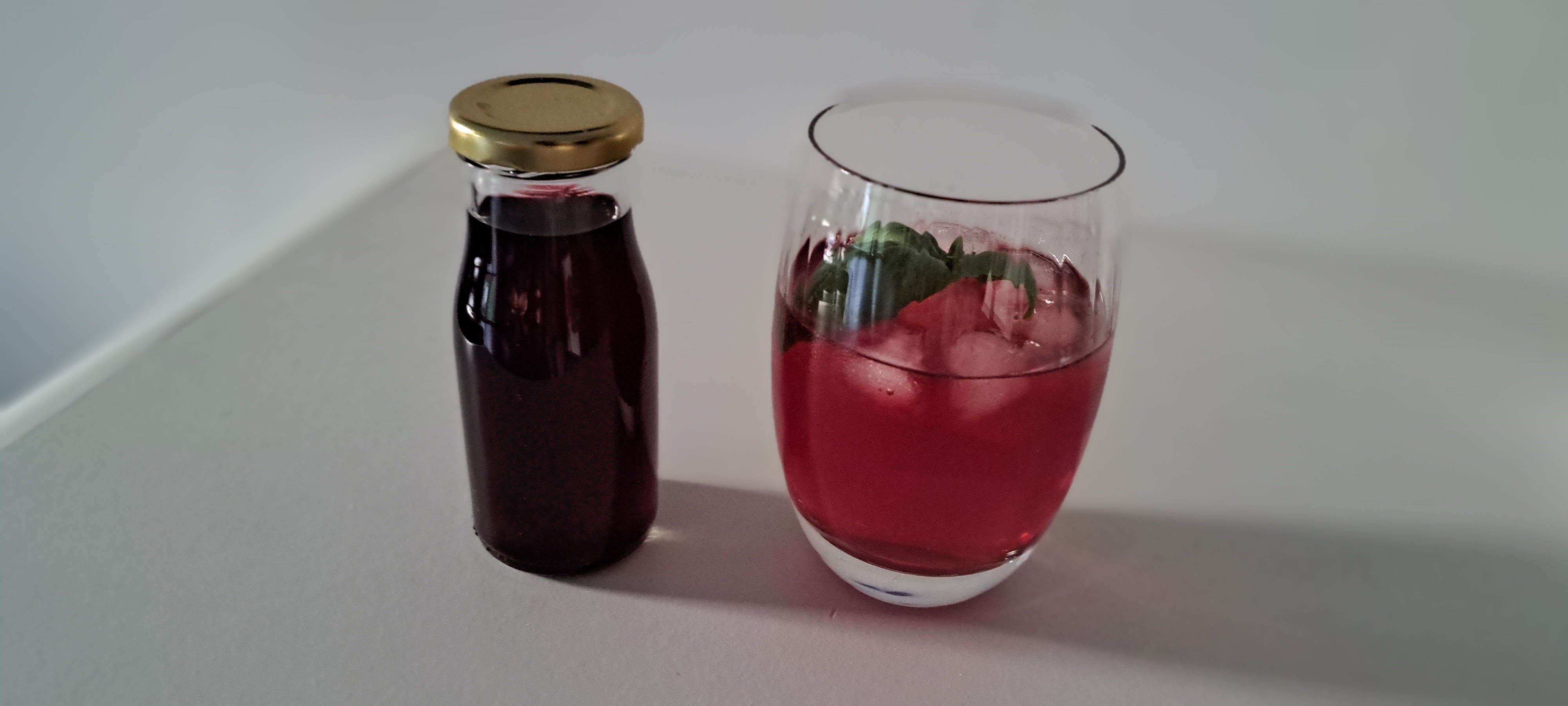 A glass containing elderberry gin with ice and a sprig of fresh basil, beside a tiny glass bottle full of dark elderberry gin.