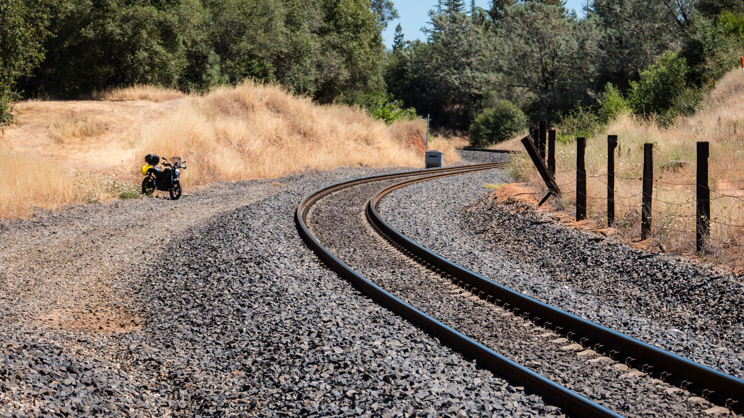 A railroad track curves right and then left as it climbs a grade through tall grasses the color of hay and towards green trees. A cattle fence with stout, wooden posts and three rows of barbed wire lines the railroad on the right. On the left, a motorcycle sits on the verge of the gravel railroad bed.