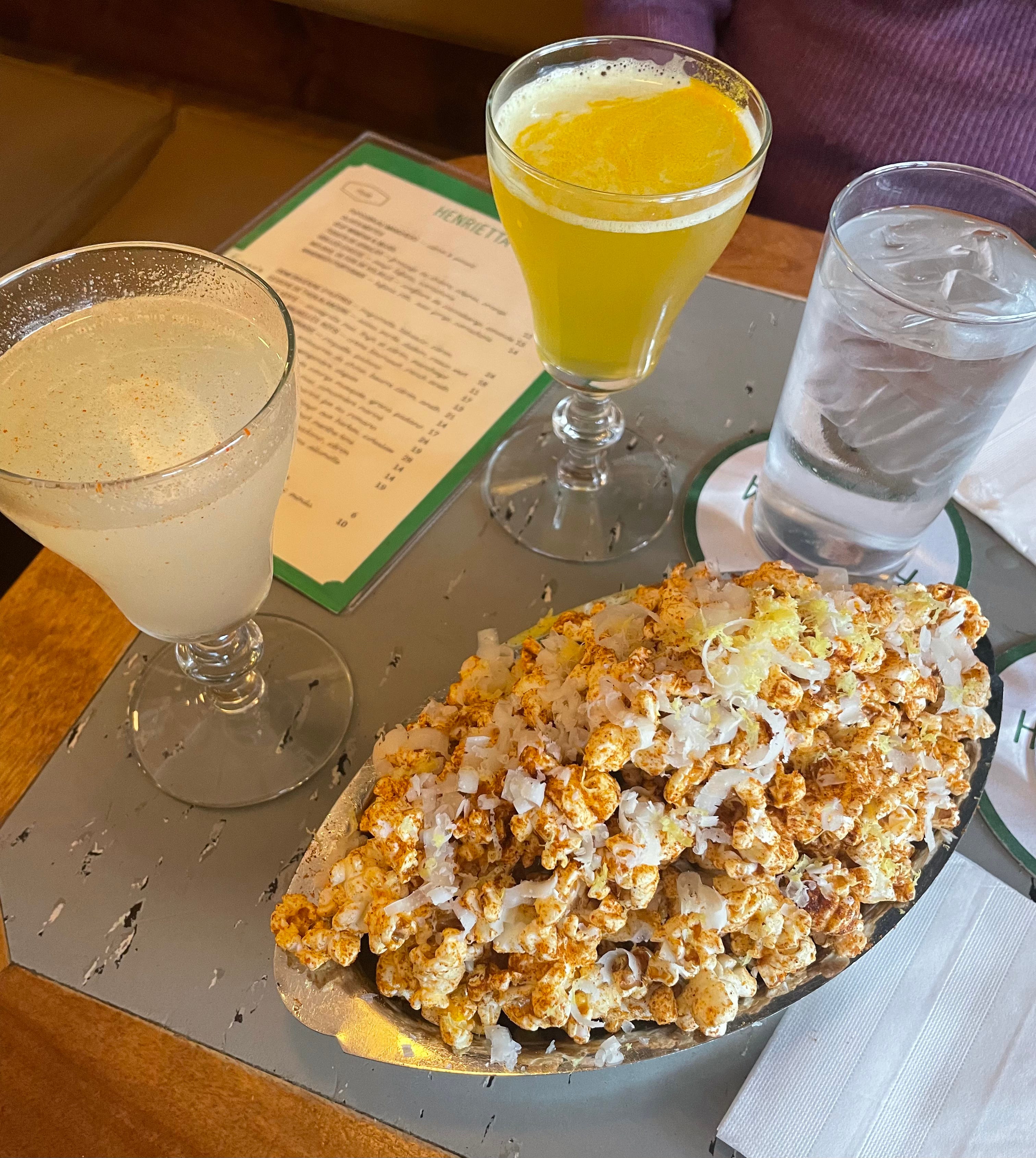 Two cocktails and a bowl of seasoned popcorn with flakes of manchego cheese all over it