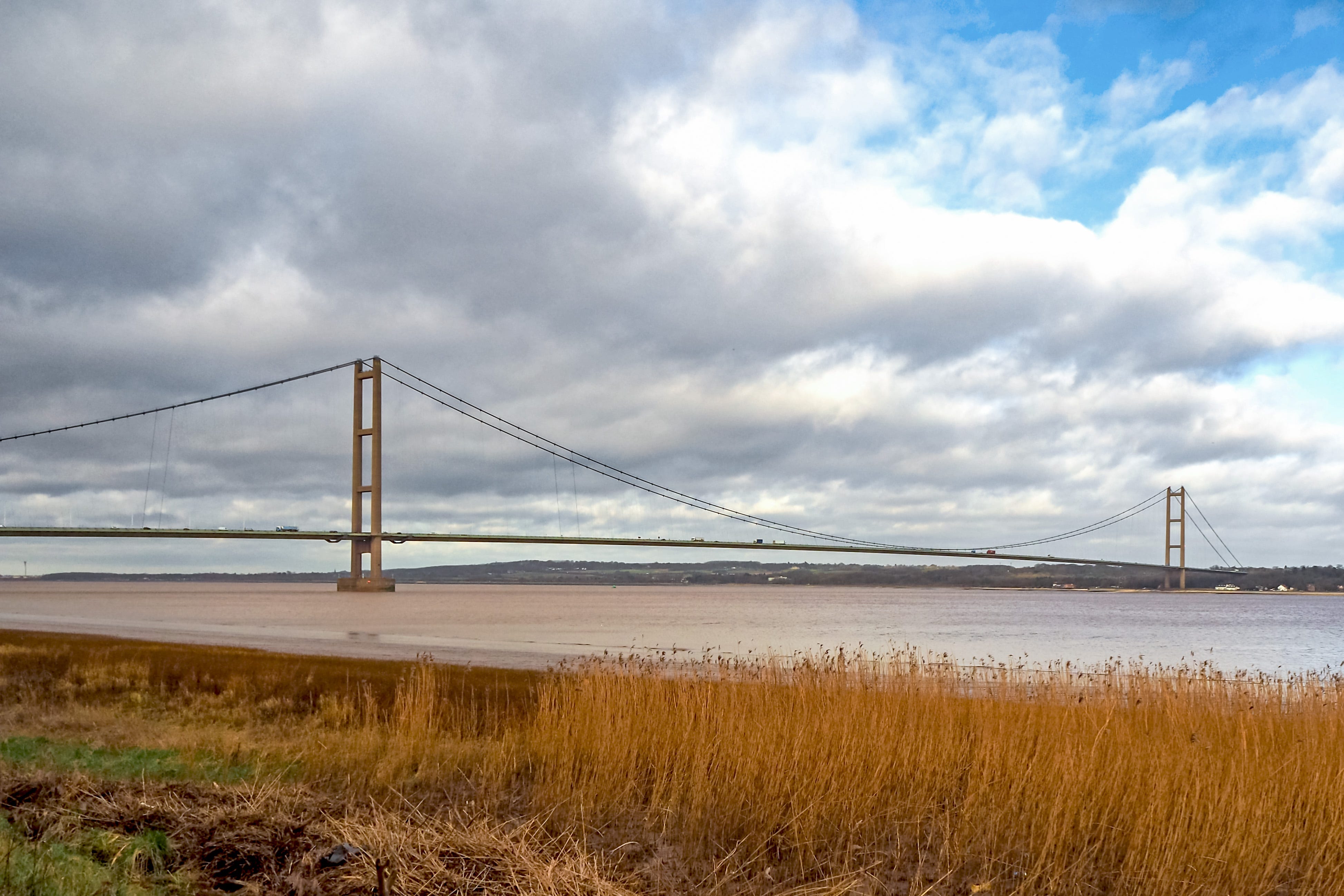 Photograph of the Humber Bridge spanning the Humber Estuary. There are reeds in the foreground of the photo, and a dramatic cloudy sky above.