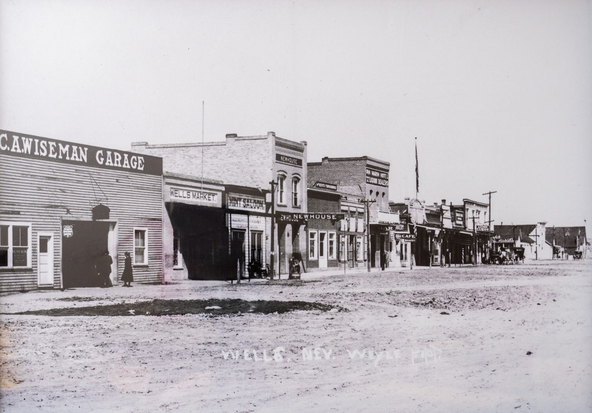 Old black and white photo of a street in Wells, Nevada. The wide street is dirt, some areas worn by the traffic of horse-drawn carriages. Alongside the road is a row of old buildings - wooden C.A. Wiseman Garage with large garage door and various windows, the brick Wells Market, with an awning over the sidewalk, the wooden Mint Saloon with the words painted right on the wooden clapboard, the two-story brick Newhouse building with the building name set high on the second floor just beneath the cornice, and several more brick and wooden buildings, most one-story, some two stories. There are electrical poles along the sidewalk and a flagpole with flag partway down the street. There are a couple of horse-drawn carriages at the end of the block. A couple of people stand outside C.A. Wiseman Garage, a man in a suit and woman in a long dark coat that reaches to her calves.