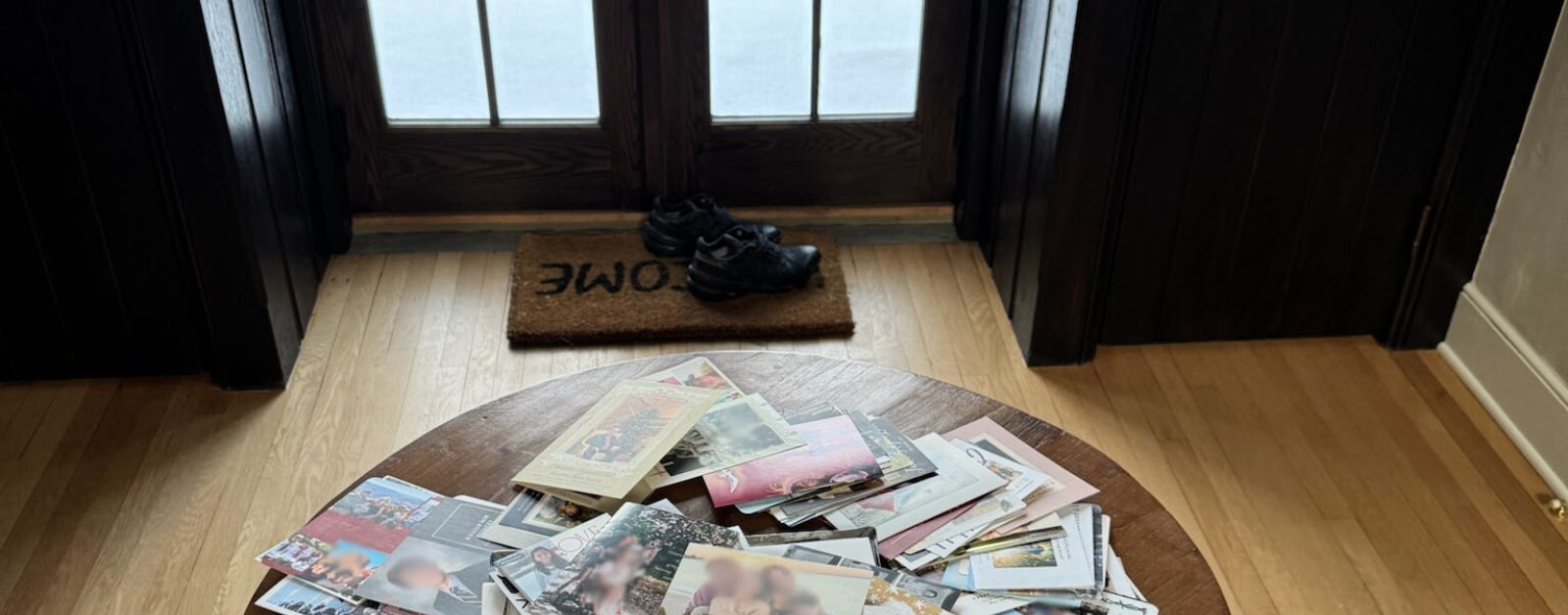 A round wooden table in the center of a room holds a large pile of holiday cards, some overlapping and scattered. The background features a set of dark wooden doors with glass panes, showing a snowy outdoor scene with a wooden deck and chairs. A doormat with the word "HOME" is visible near the door, along with a pair of black shoes. 