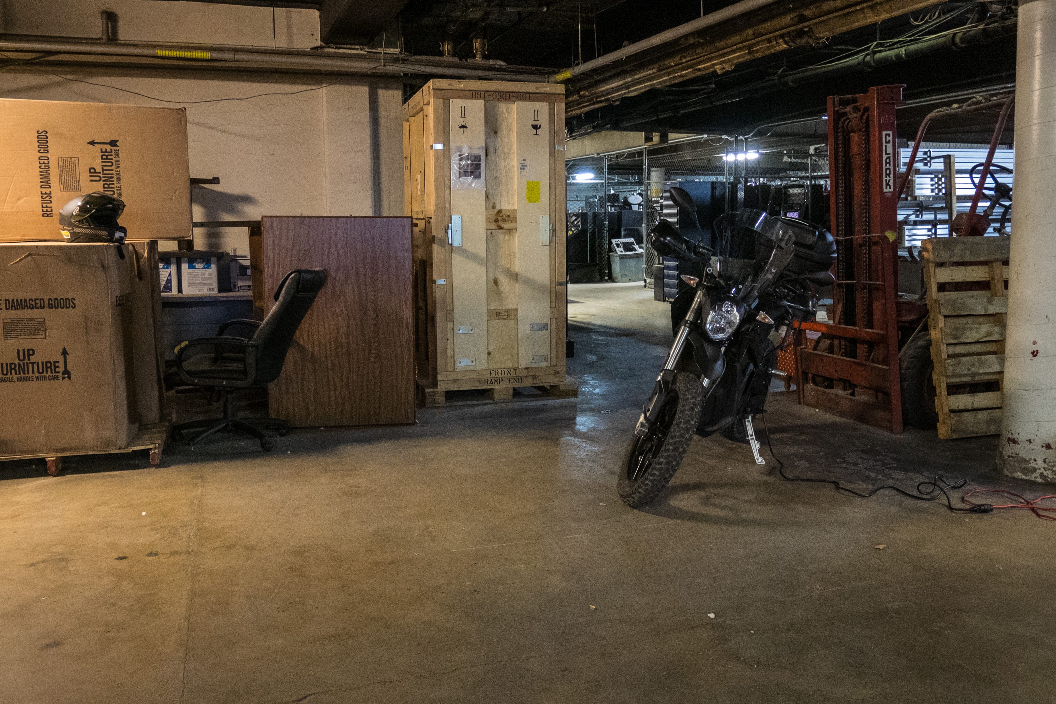 An electric motorcycle sits parked in a low-ceilinged storage area, an extension cord running from the motorcycle off to the right edge of the photo. Around the motorcycle are random objects - an orange forklift behind the motorcycle, a tall, sturdy crate with fragile icons on it to the left of the bike, old wood-veneer office furniture and an office chair besides that, and large cardboard boxes with the words, "furniture / UP" with corresponding arrows. One of the boxes is laying on it's side. One of the boxes has a motorcycle helmet on it. Beyond, are shipping palettes and various other mechanical objects.