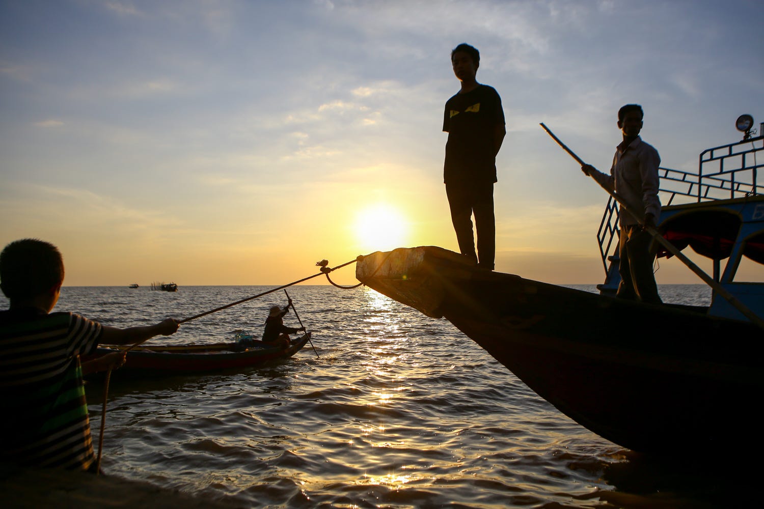 A photo of a long-tail boat with two men standing on it, silhoutted against the setting sun. The lake is flat, with a few smaller boats in the background. A boy holds a rope from the boat in the frame, helping bring it to the floating restaurant. 