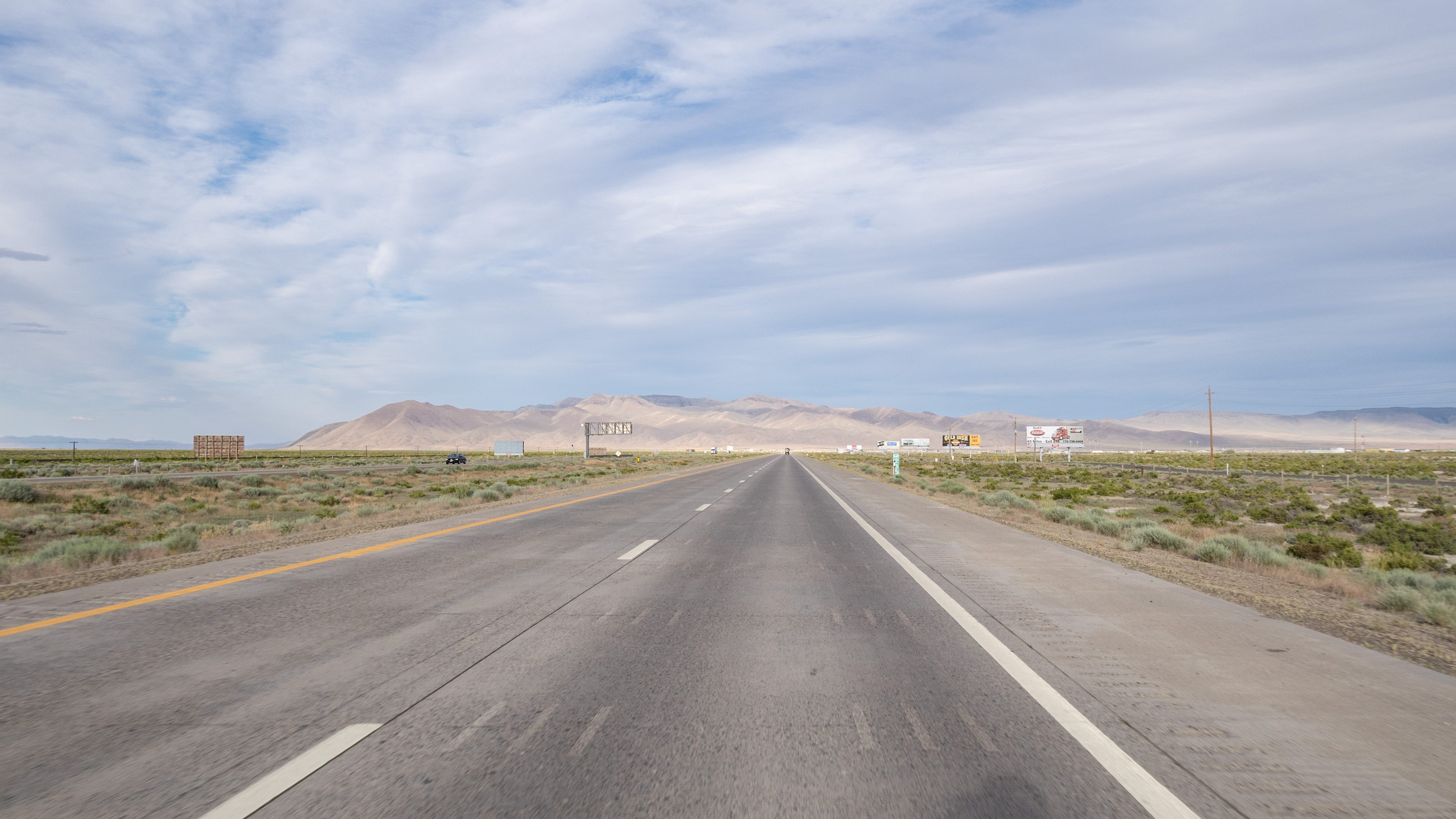 Photo of Intersate 80, heading east. The two eastbound lanes (and broad shoulder) are flat and empty. On either side of the highway is the desert and scrub brush. In the distance, large billboards line the side of the road. Further, beyond the vanishing point of the highway, is a wall of naked, treeless, rugged mountains. The sky is blue and streaked with clouds. The sun is high overhead.