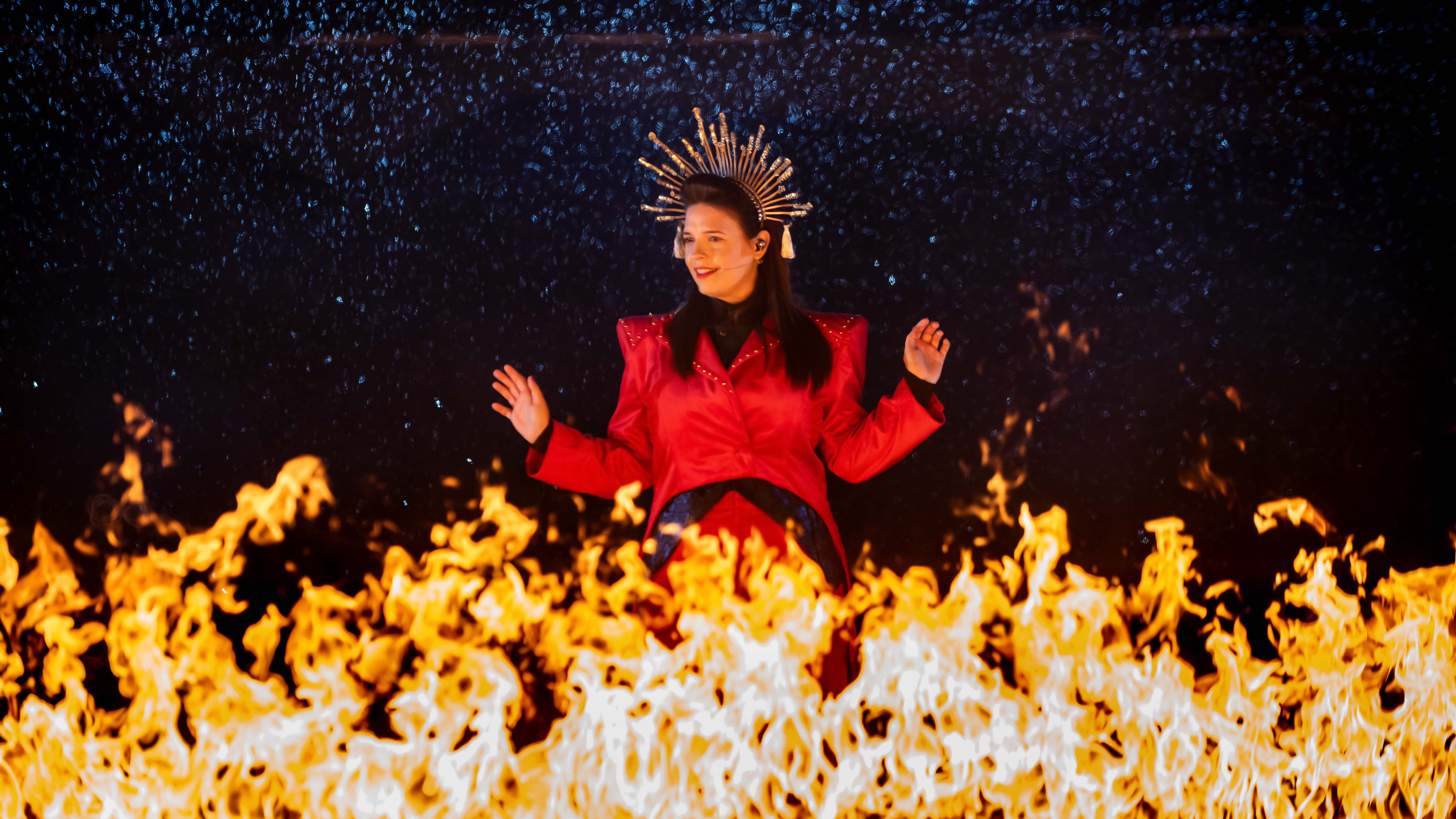 A young woman wearing a red jacket and an angel-like headress rises above the flames.
