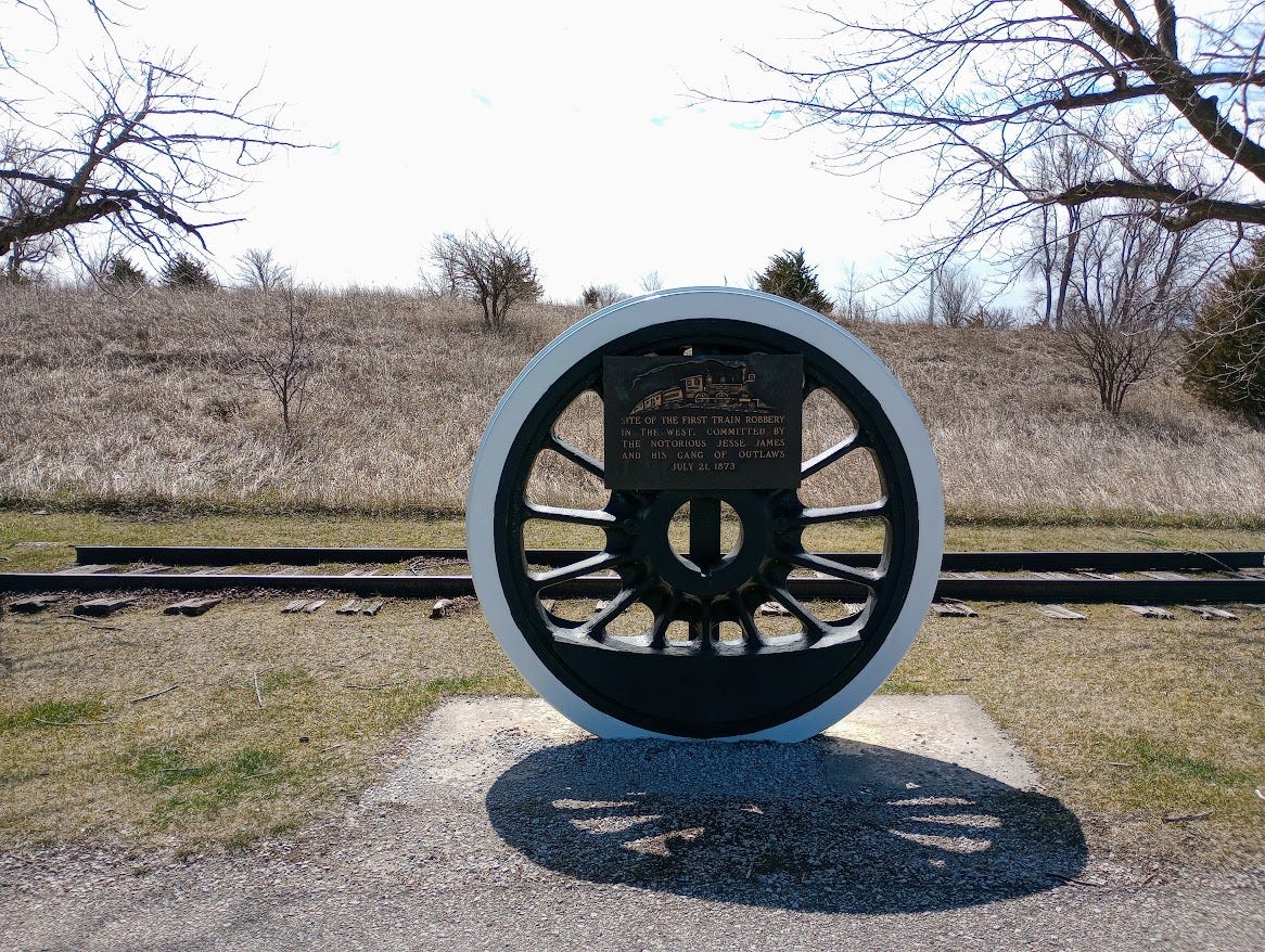 Monument in shape of railroad wheel with small section of track behind set in grasses with trees