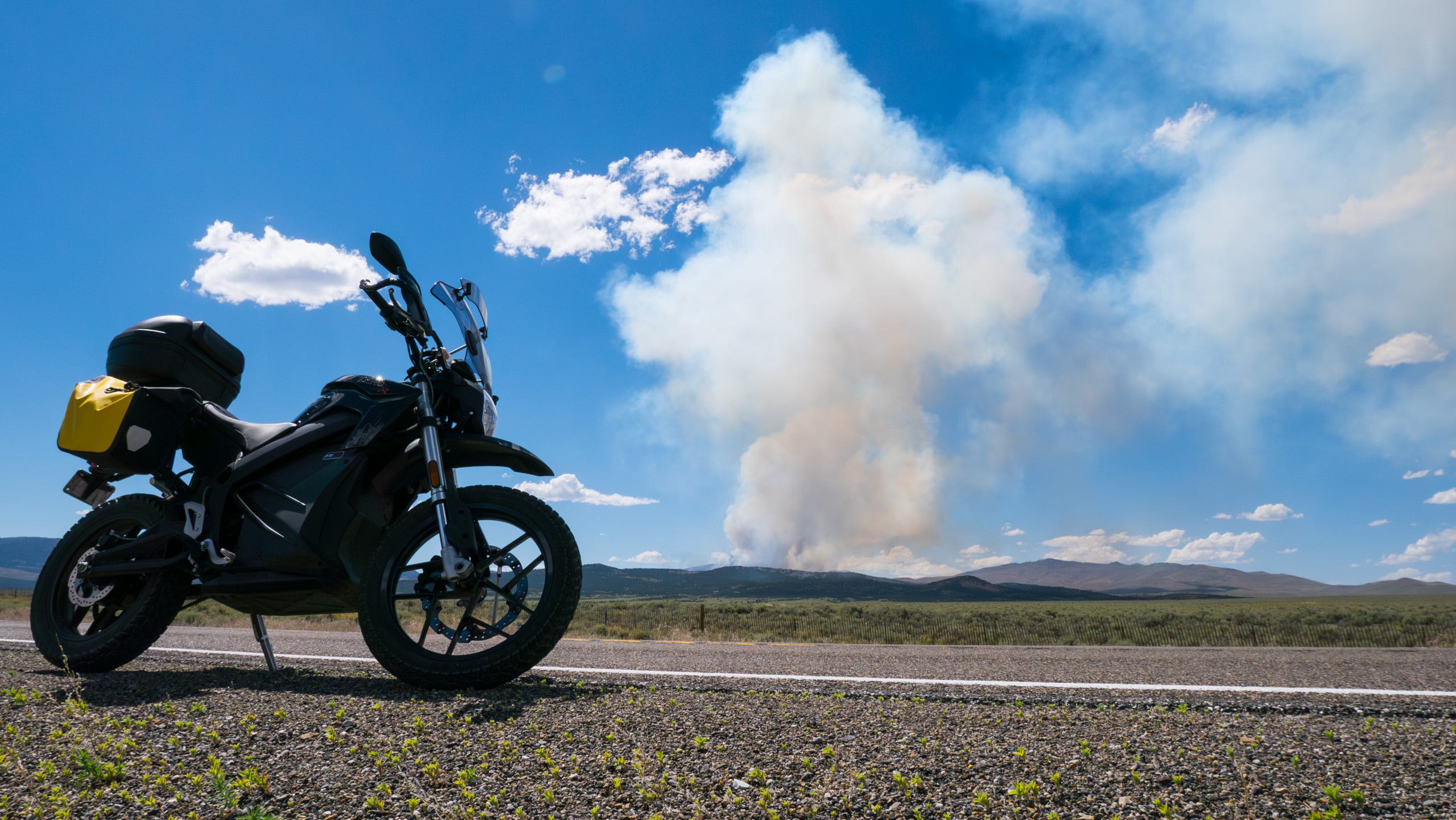 Photo of an electric motorcycle in the foreground parked on the side of the road with large plumes of smoke filling the sky from a forest fire in the distant mountains.