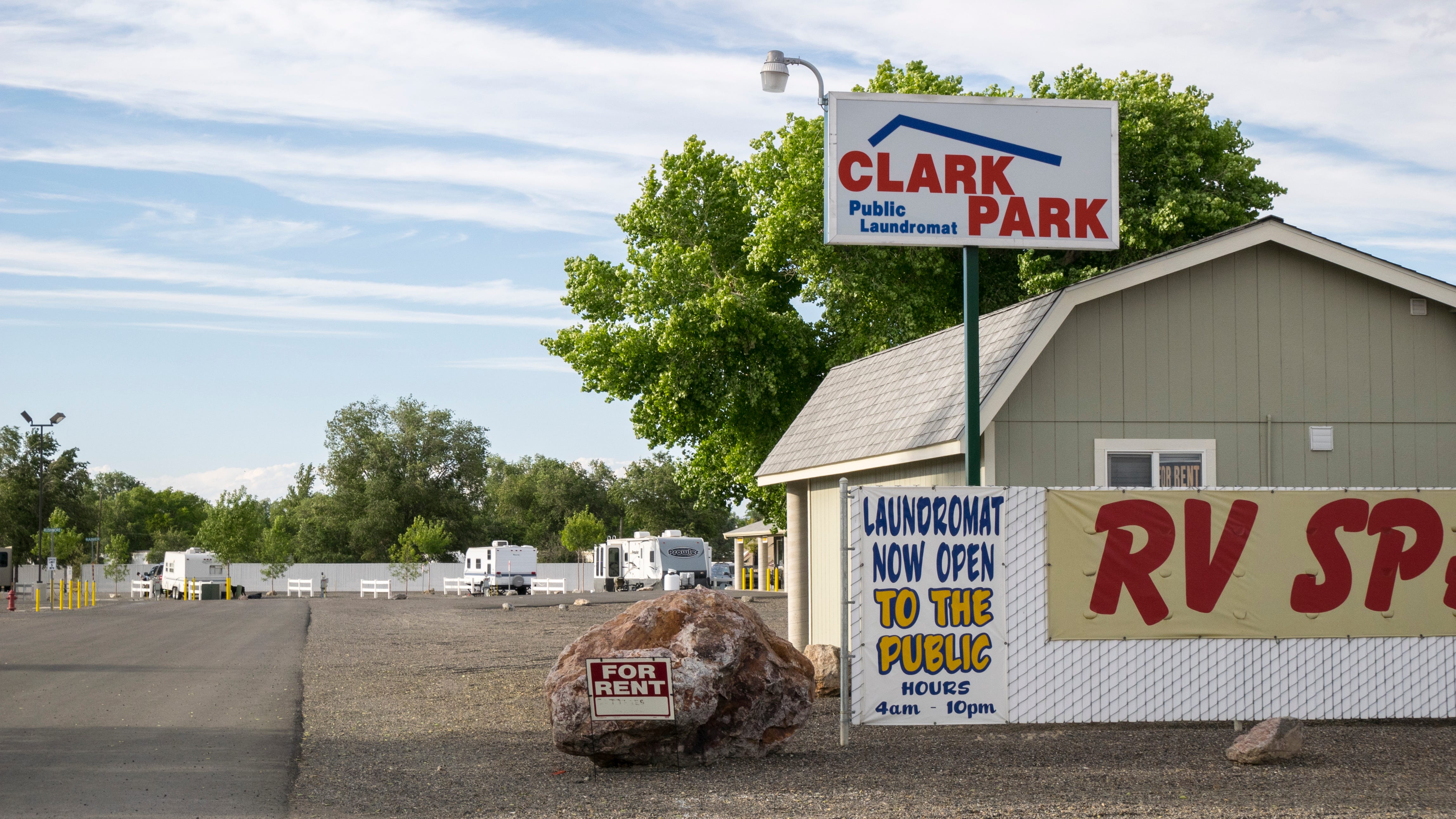 Photograph at the entrance to Clark Park RV park. A wide asphalt road leads into the park. The rest of the surface is gravel. In the background are a handful of white RV campers. A large red boulder sits at the entrance with a "For Rent" sign on it. Beside the boulder is a simple chainlink fence with white slats. A sign "Laundromat Now Open to the Public / Hours 4am - 10pm" is hung on the fence. Another sign, incomplete, hangs on the fence; it reads "RV SP..."   Behind, on a metal pole, is a backlit signt "Clark Park / Public Laundromat" Just behind the fence is a small barn-like building with a Mansard roof. The place feels empty.