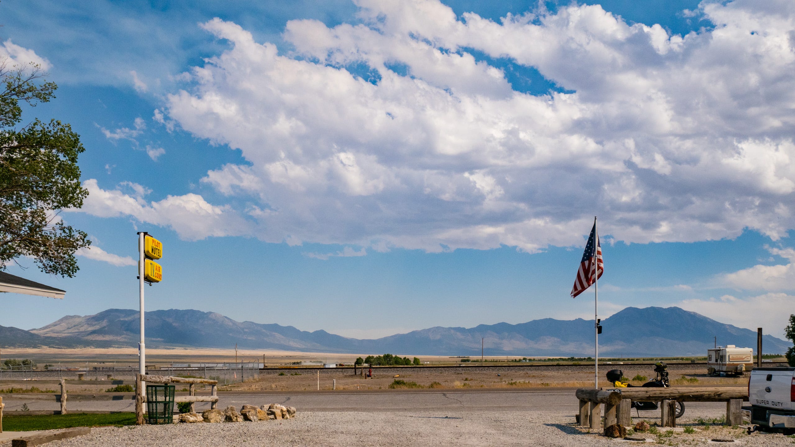 Photo looking from the parking area of a small hotel, across the road, across the railroad tracks, across a broad valley towards distant, jagged mountains in neighboring Utah. Above the mountains, the sky is filled with puffy clouds in a blue sky. Along the roadside is a wooden, split rail fence on the left, a yellow motel sign on a white pole. On the right, is the US flag on a flagpole. The composition is balanced and serene.