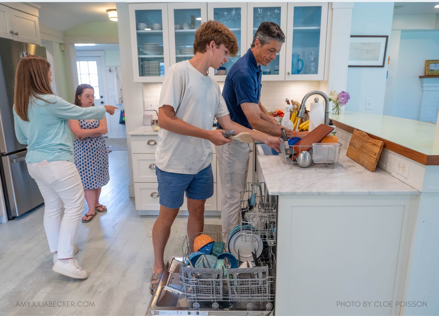 Amy Julia's family working together in a kitchen: Peter and William unload the dishwasher, while Amy Julia and Penny are in the background.