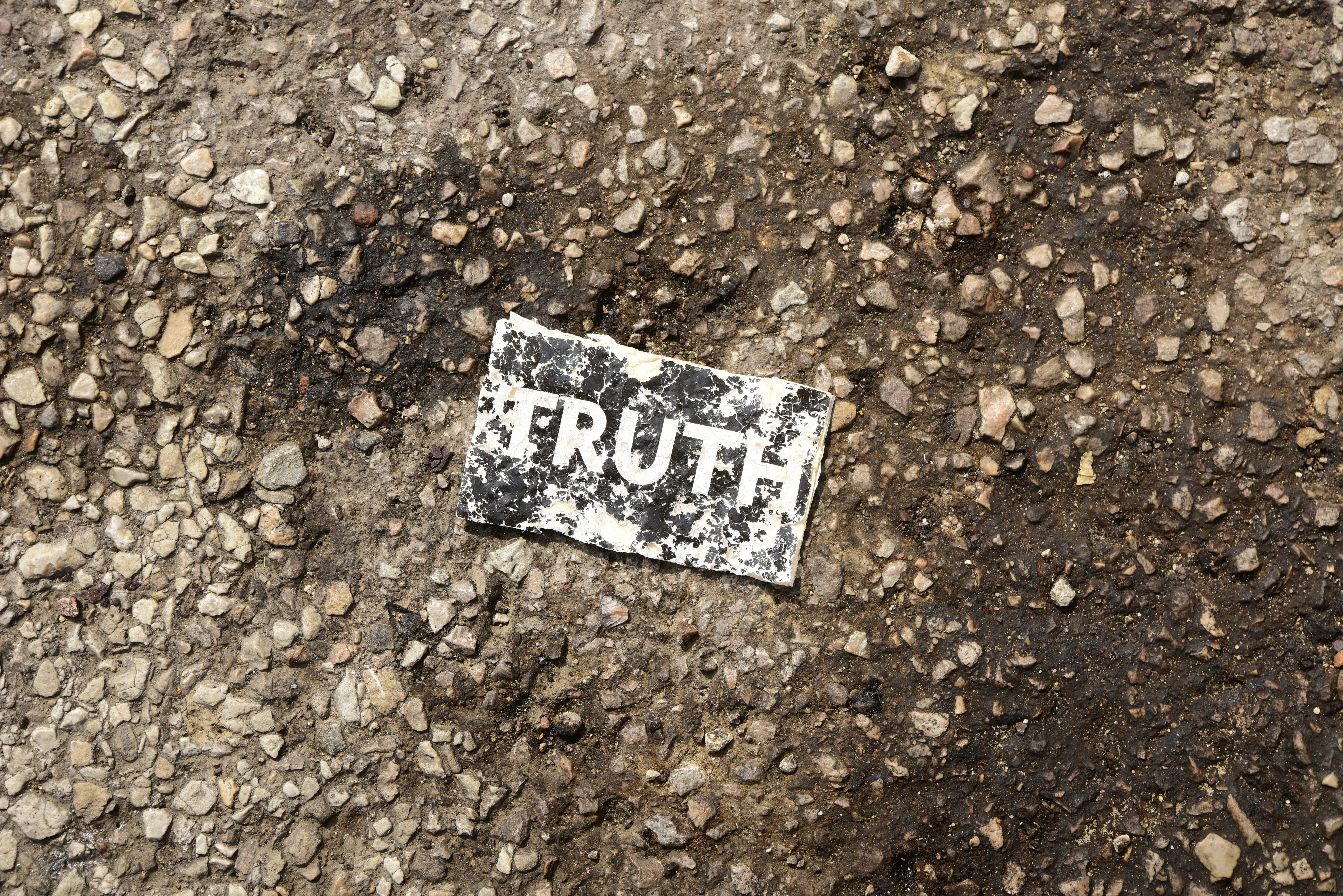 A piece of worn out paper. It is black. The word Truth is written in white. It is laying on a rough pebbled ground that is a mix of greys and browns