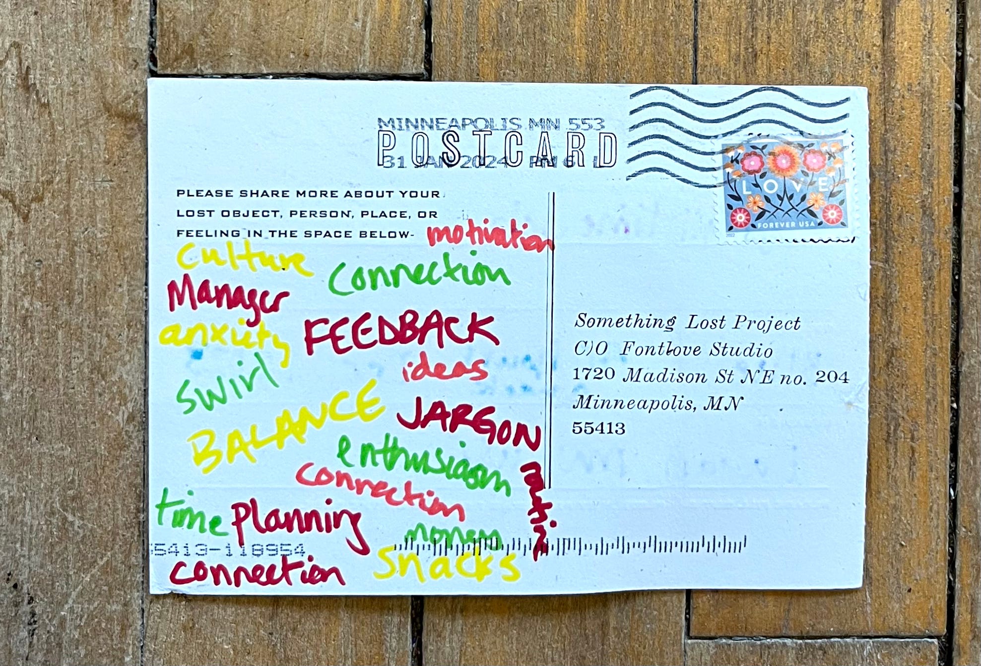 The back of a postcard with many single words hand-written in different colors of markers. The words are jumbled and include motivation, culture, connection, anxiety, feedback, ideas, enthusiasm, swirl, jargon, routine, money, and snacks.
