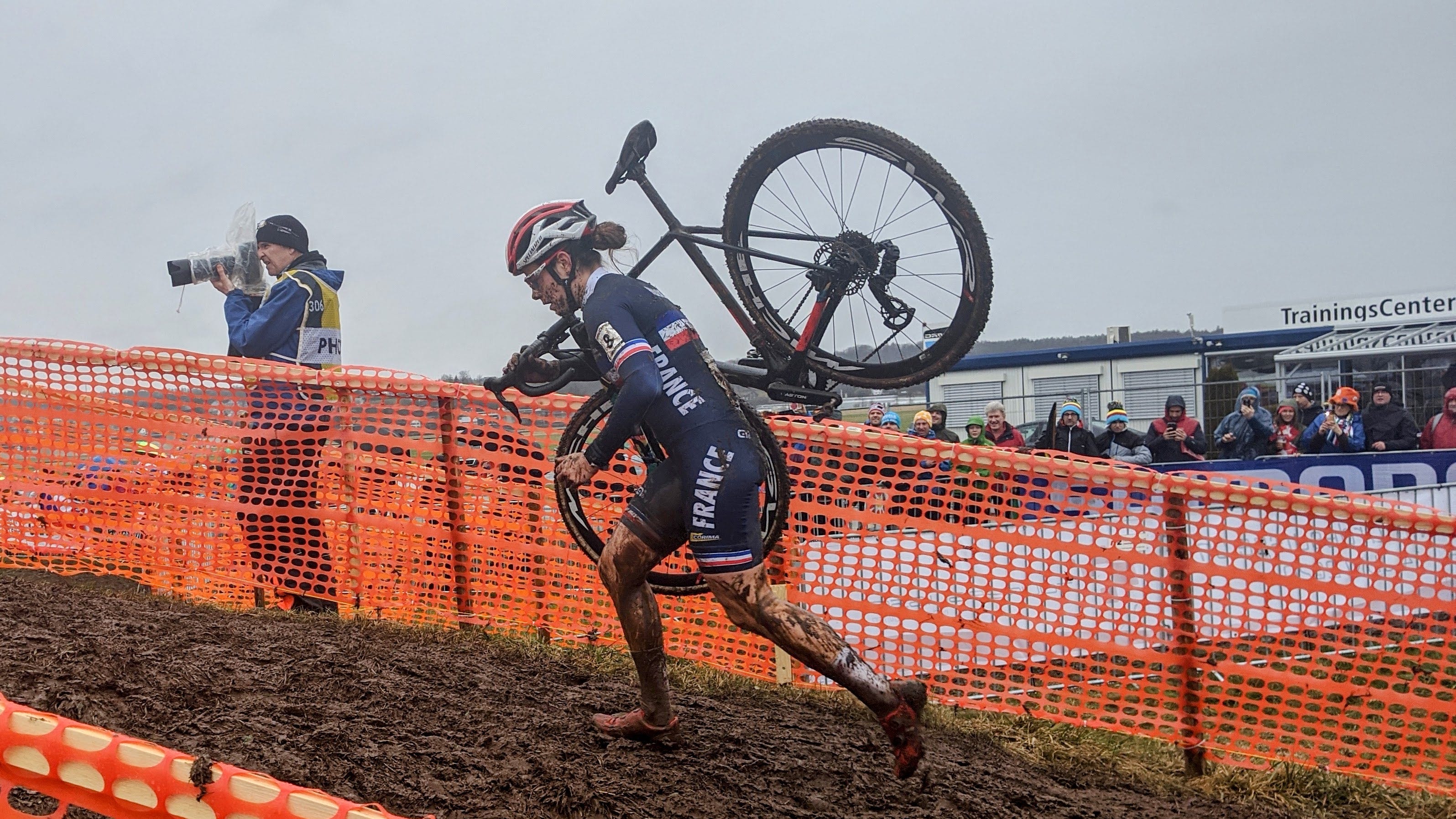 A female cyclist runs up a muddy hill, carrying her bicycle on her right shoulder at a cyclo-cross race. There is an orange plastic fence separating her from the crowd