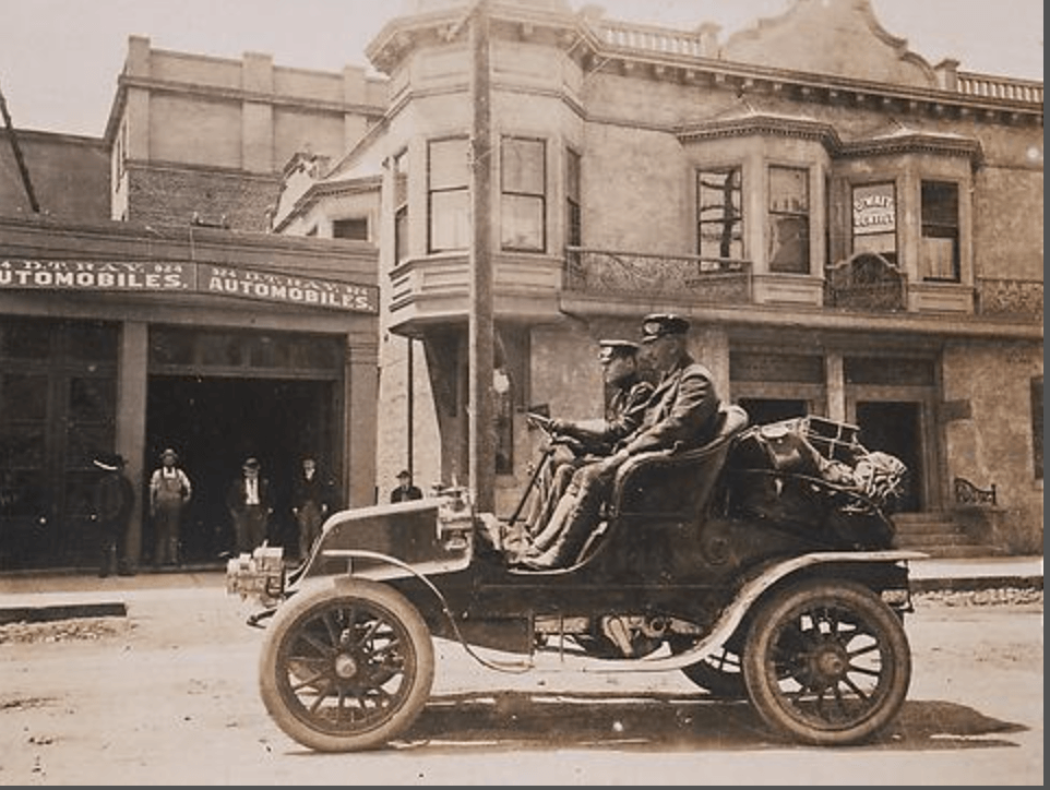 Old sepia-toned photo of Dr. Horatio Nelson Jackson and Sewall K. Crocker driving a Winton automobile down an urban street. The Winton features four tall and narrow wood-spoked wheels at the corners and a simple, roofless, two-person passenger area. There is a short sloping bonnet up front and a sloping rear bonnet in back, upon which has been strapped various boxes and packages for the trip. A pair of metal headlights sit at the front of the car. A simple fender runs from the front wheel, curves down to become a running board, and then curves up again over the rear wheel. Jackson and Crocker are wearing matching outfits - slim pants/leggings, boots, sturdy jackets, and flat-topped driver's caps. The younger of the two men (presumably Crocker) is at the steering wheel, which is nearly horizontal and sitting nearly in his lap as the thin steering column descends from the steering wheel nearly vertically to the floor. There are no doors or windows and some of the mechanical parts of the car are visible below the carriage. Across the street is a two-story stone building with bay windows and a cornice. Besides that is a tall one store garage, with what appears to be two tall garage doors. One of the doors is open, and four men stand at the door watching the car drive by. One is wearing overalls (likely a mechanic) while the others are in jackets and trousers and hats. The street is unpaved.
