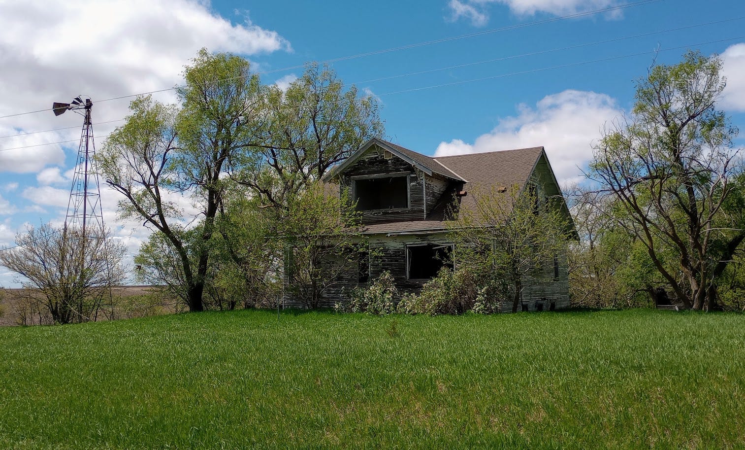 House with decaying boards, missing windows, but relatively new roof, next to small windmill and trees