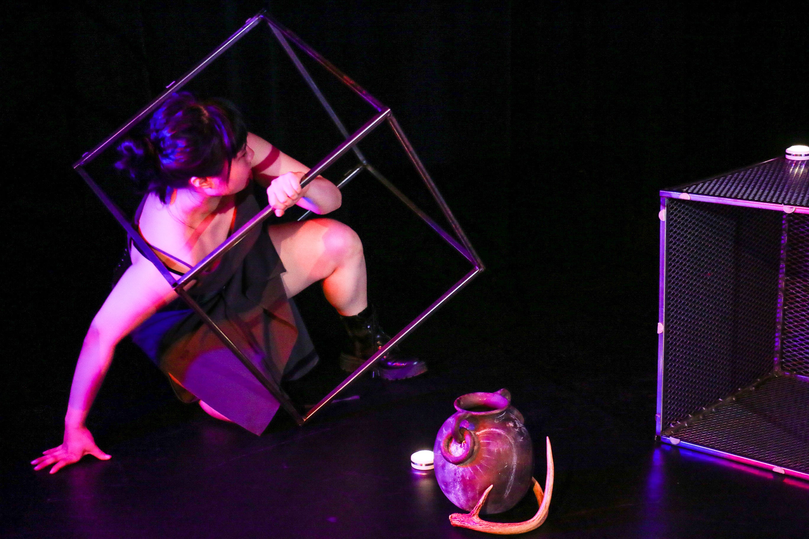 A young woman, Yitong Chen, is off-center in a metal frame of a box. Other objects are in the space including a mesh metal box, solar lighting elements, a dear antler and a ceramic jug.