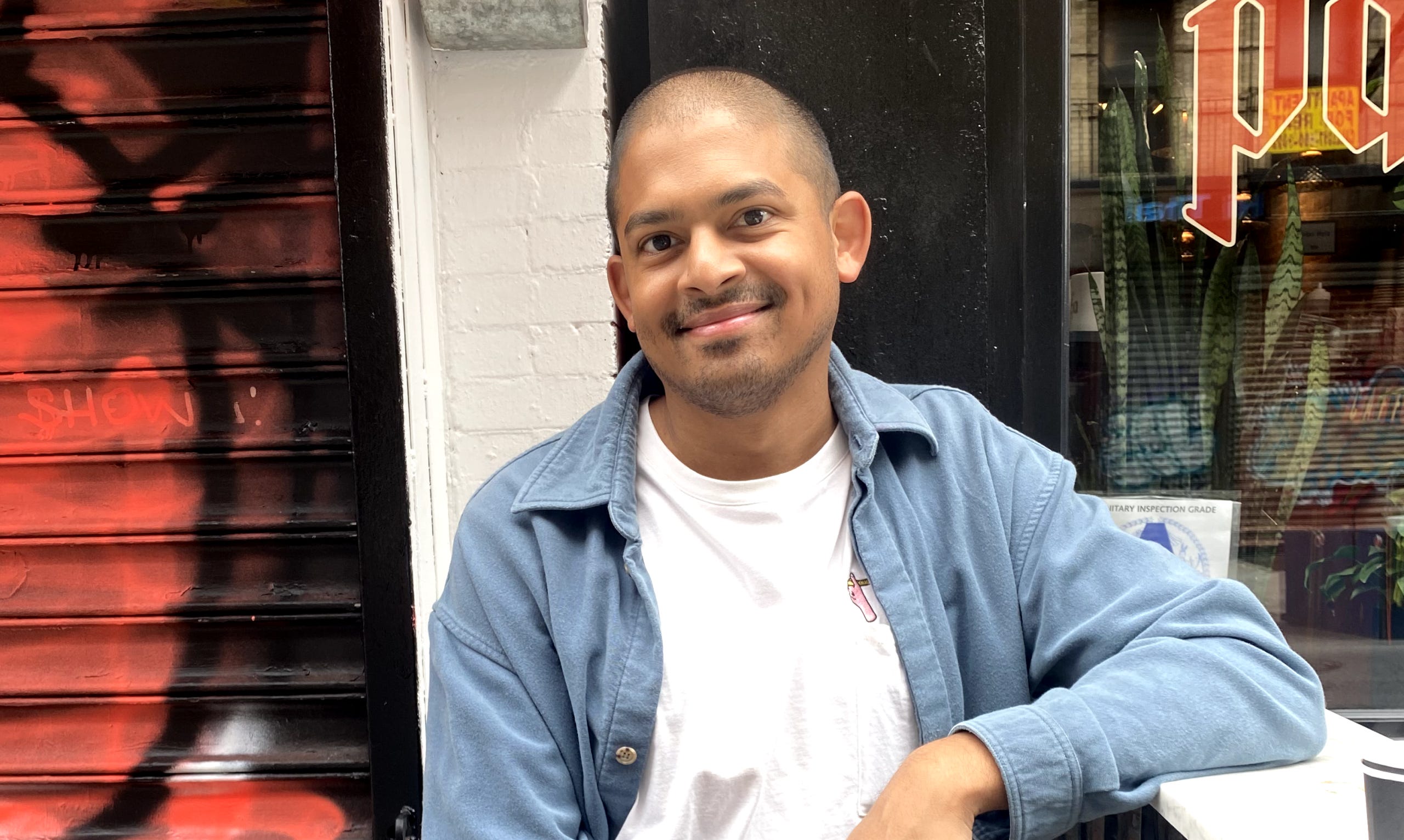 Kirk Fernandes sits and poses smiling at the camera in a blue jacket over a white shirt in front of an outdoor space.