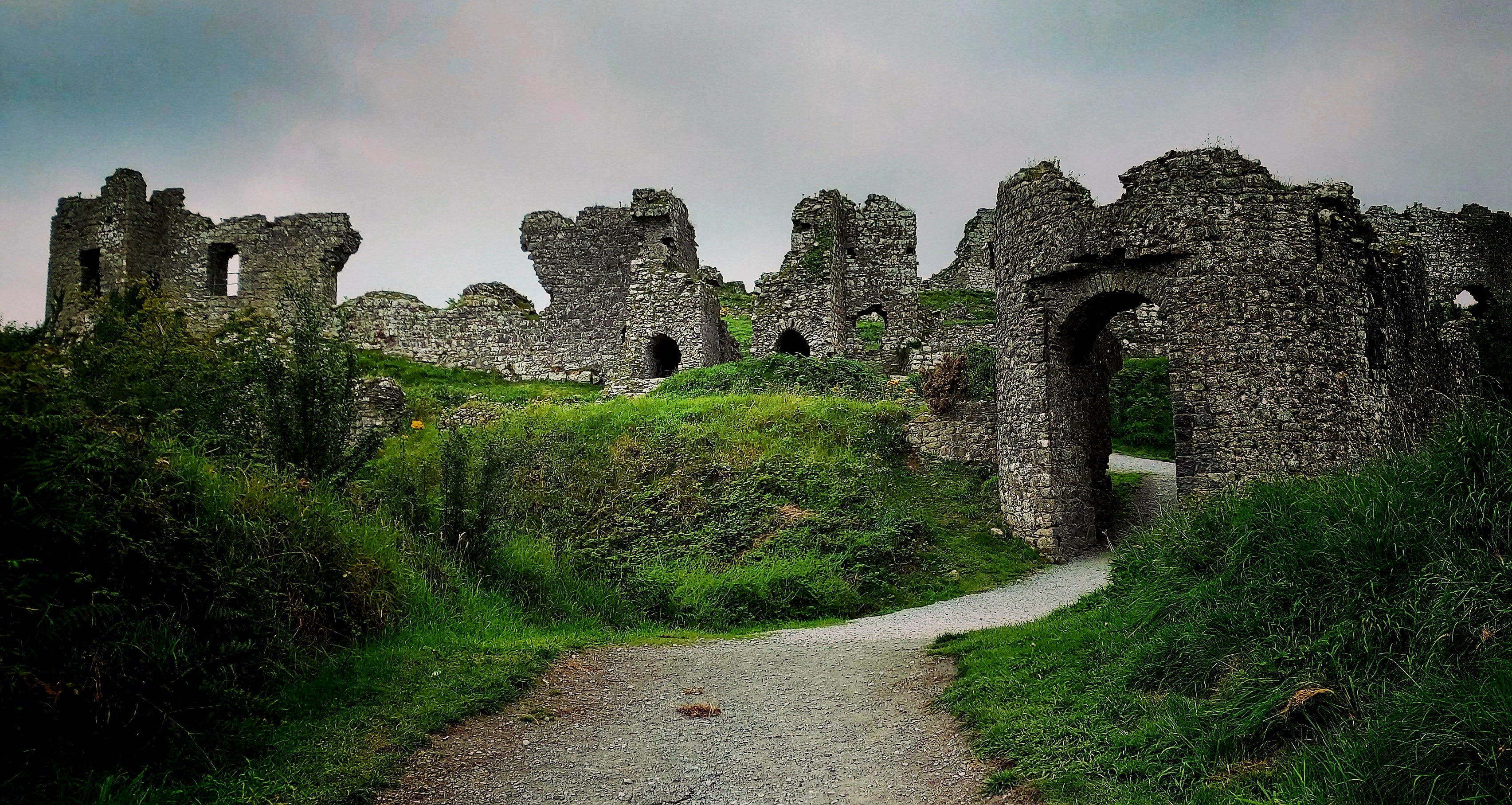 Image of tumbled stone castle silhouetted against a cloudy skyline, stonework is grey, hillside is green and grassy, there is a fairy-tale atmosphere in this image.