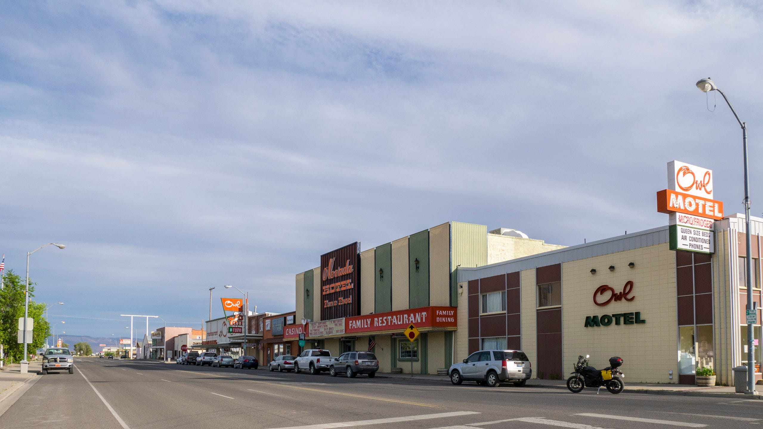 Photograph of downtown Battle Mountain. The main street is wide - two lanes and parking in each direction, and the building are low one- and two- story buildings. The Zero motorycle is parked across the street in front of the rectilinear, mid-century Owl Motel with a simply pale yellow concrete block facade interrupted by vertical areas of maroon panels trimmed by thin aluminum mullions. The mullions create a grid within which simple rectangular window site. An old neon sign "Owl Motel / Microfridges / Queen Size Beds / Air Conditioner / Phones" juts out from the facade. The name "Owl Motel" is set upon the facade, "Owl" in script, "Motel" in all caps. Besides the Owl Motel is a slightly taller building, the "Nevada Hotel", with vertical panels of pale yellow and green and a red marquee that reads "Casino" and "Family Restaurant." Beyond, are low one story retail stores. The street sits low in the photo, beneath a blue sky streaked with clouds. The overall vibe is quiet.
