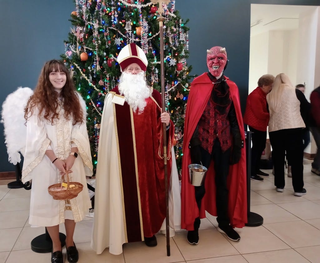 Woman dressed as angel, man dressed as St. Nicholas, and man dressed as Devil standing in front of Chritmas tree