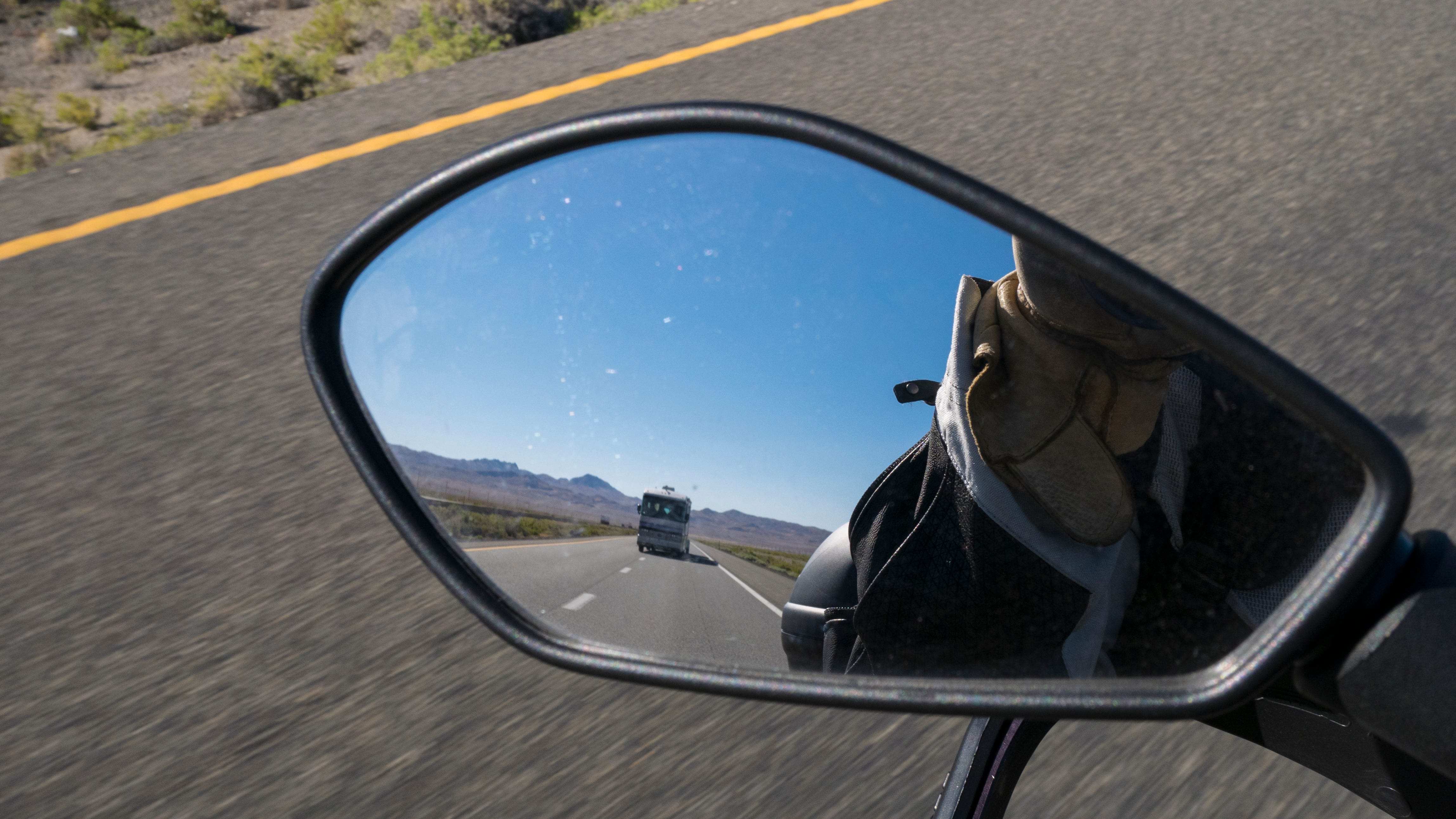 Photo from the seat of a moving motorcycle on the interestate. The photo is of the motorcycle's left rearview mirror. In the mirror is a large RV behind the motorcycle switching lanes to pass the motorcycle. Beyond the RV are rugged brown mountains. The mirror also shows the rider's gloved hand and a portion of his jacket. Below the mirror, the asphalt of the interstate is blurred by the motorcycle's speed.