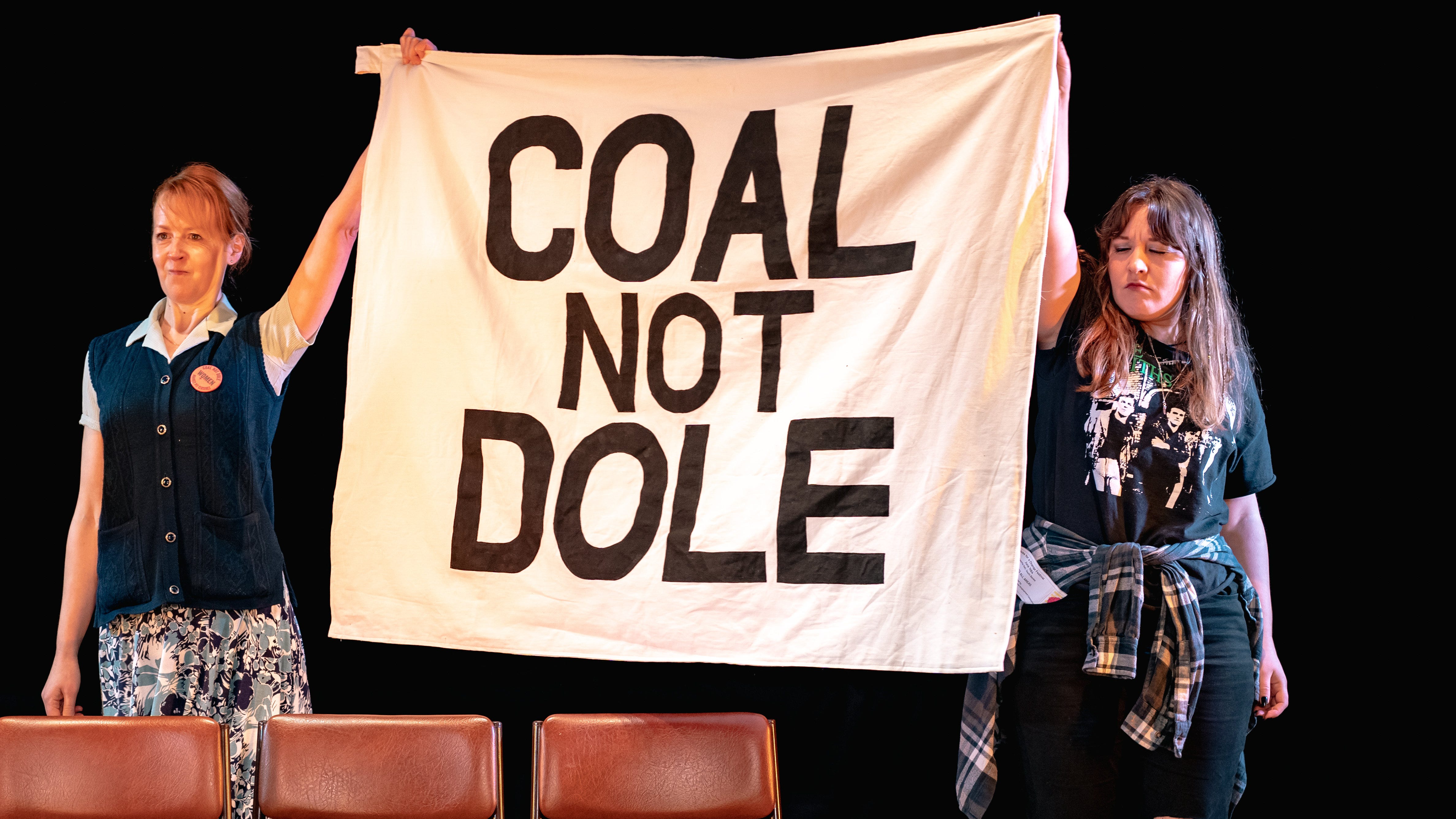 Two women dressed in 1980s clothing hold up a banner saying Coal Not Dole.