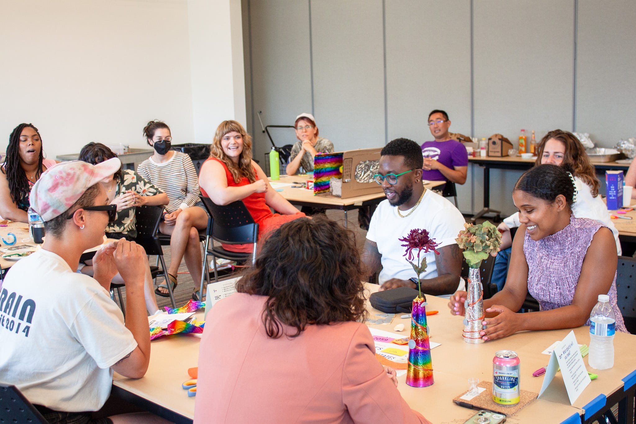 In this wide shot of a room full of workshop participants, about 10 people look on, smiling, while listening to MARS (a brown-skinned person in a white shirt and glasses) describe his creation. Imagined abolitionist artifacts populate the table tops, made out of art supplies like cardboard and flowers and more.