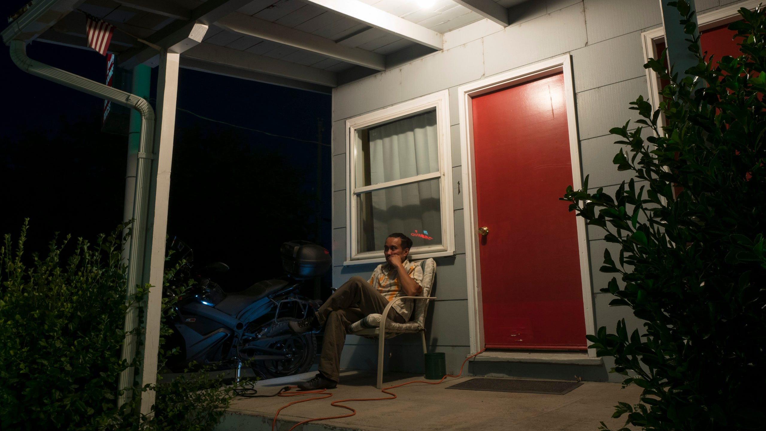 A man sits on the porch outside of his hotel room on a lawn chair. The door to his room is red. A single overhead light illuminates the concrete porch. Sneaking out of the corner of the doorway is an orange extension cord that snakes across the concrete and past the man to a black electric motorcycle sitting in the dark. A couple of shrubs frame the photo. A small US flag hangs from one of the posts holding up the overhanging roof.