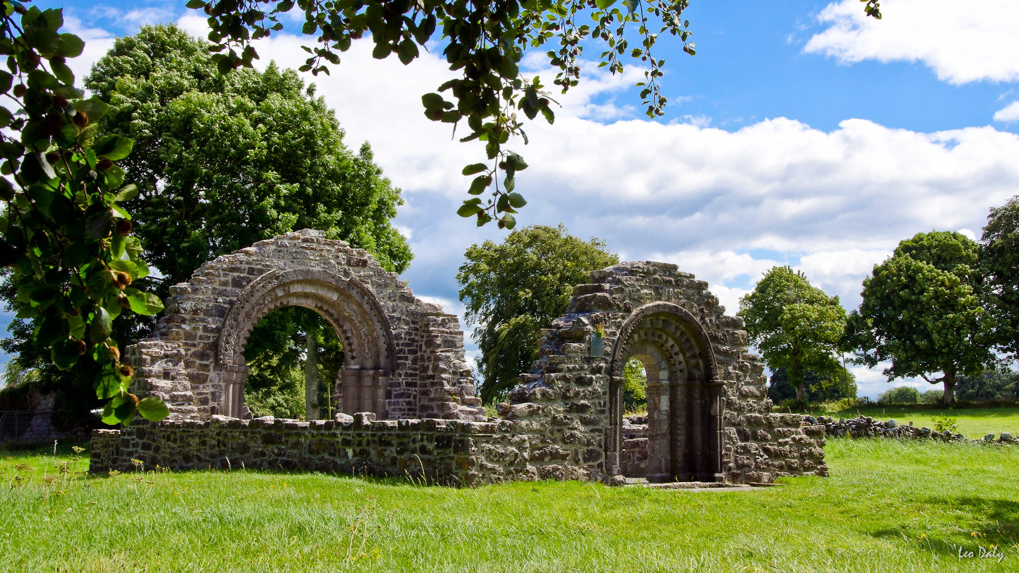 The ruined Nun's Church at Clonmacnoise, showing the two end walls of the building including finely carved arched entrance, and all that remains of the side walls. This roofless structure is situated on the edge of the main site, surrounded by its own wall and gardens, now featuring tall trees in full leaf beneath a blue sky scattered with white clouds.